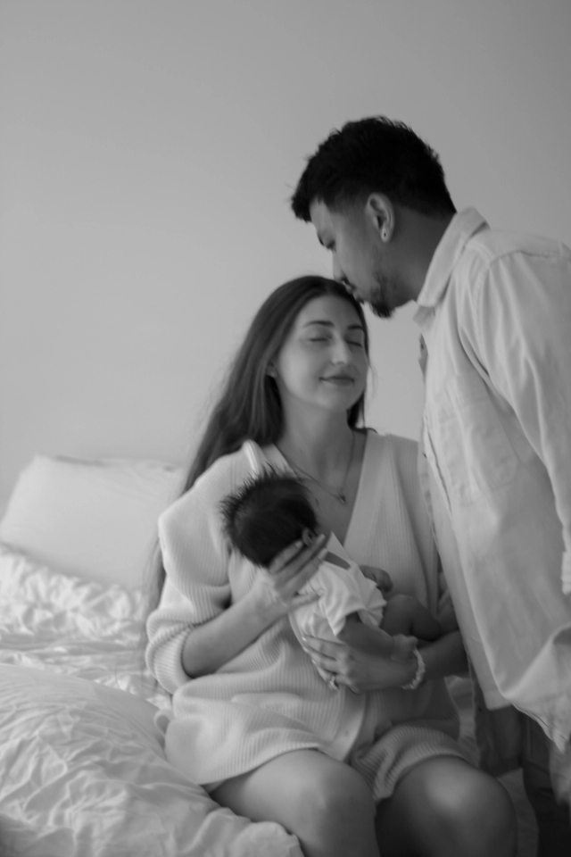 mom sitting on bed holding newborn baby while dad stands beside them kissing her forehead, Black & white - Newborn Photography