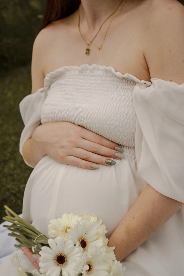 close up of mom in white dress holding her baby belly in one hand while holding white flowers in the other - Maternity Photography