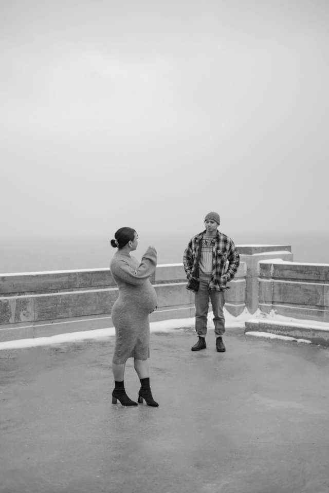 Classy winter maternity photoshoot on lakeside rooftop with first-time parents, woman is standing in foreground looking back at husband in the background. 