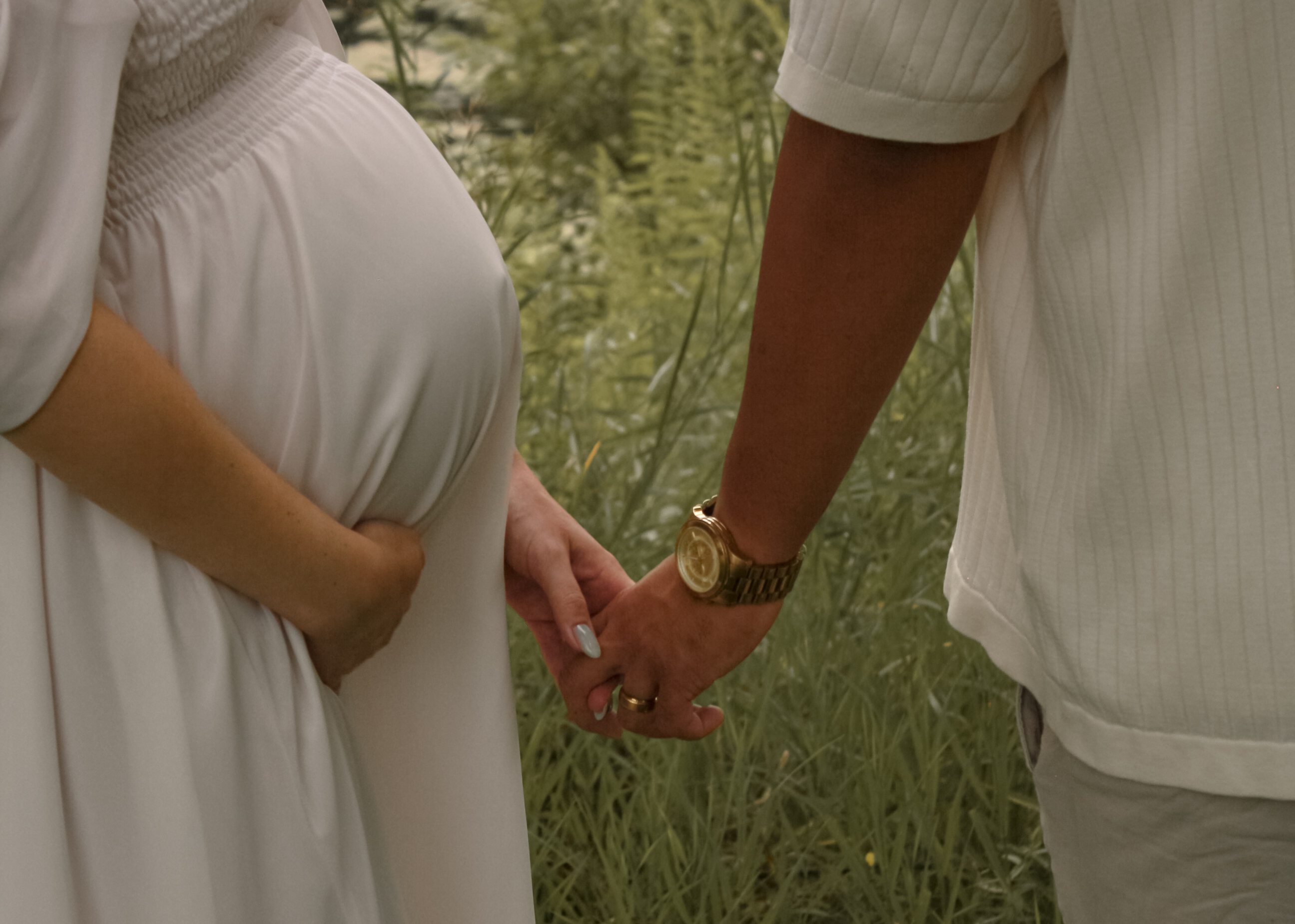 Close up of couple holding hands while the woman is holding her baby bump and man is wearing a gold watch with grass in the background - Maternity Photography