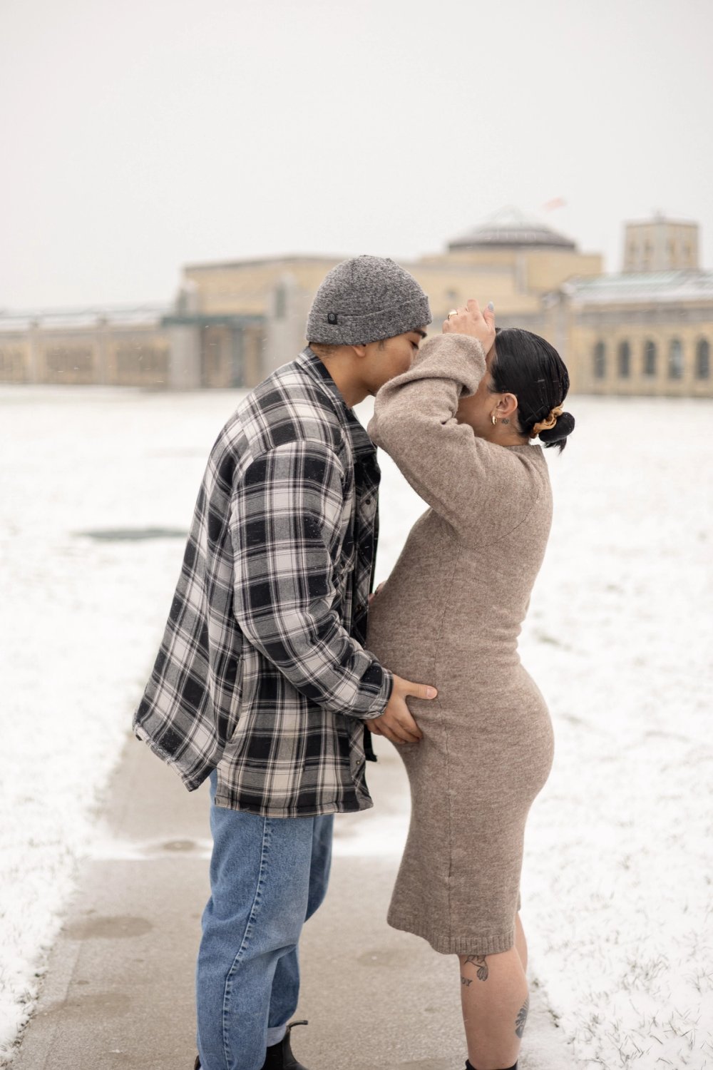 Expectant mother and husband kissing against the background of an Art Deco architecture of the R.C. Harris Water Treatment Plant in Scarborough, ON during a winter maternity photoshoot. - Newmarket Maternity Photographer