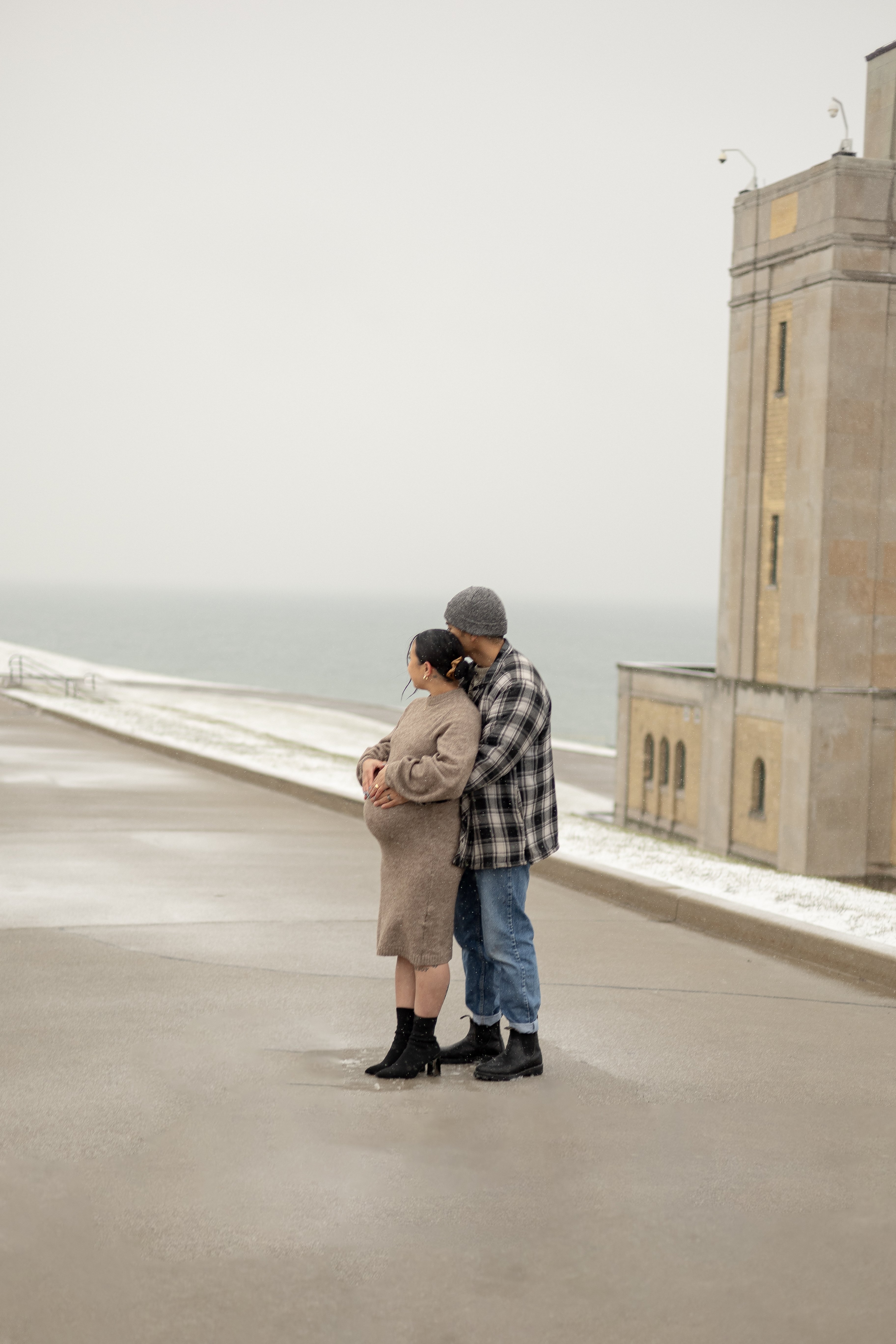 Winter maternity photos at R.C. Harris Water Treatment Plant in Scarborough, ON. Dad hugging mom from behind holding baby bump while looking out at the lake - Newmarket Maternity Photographer