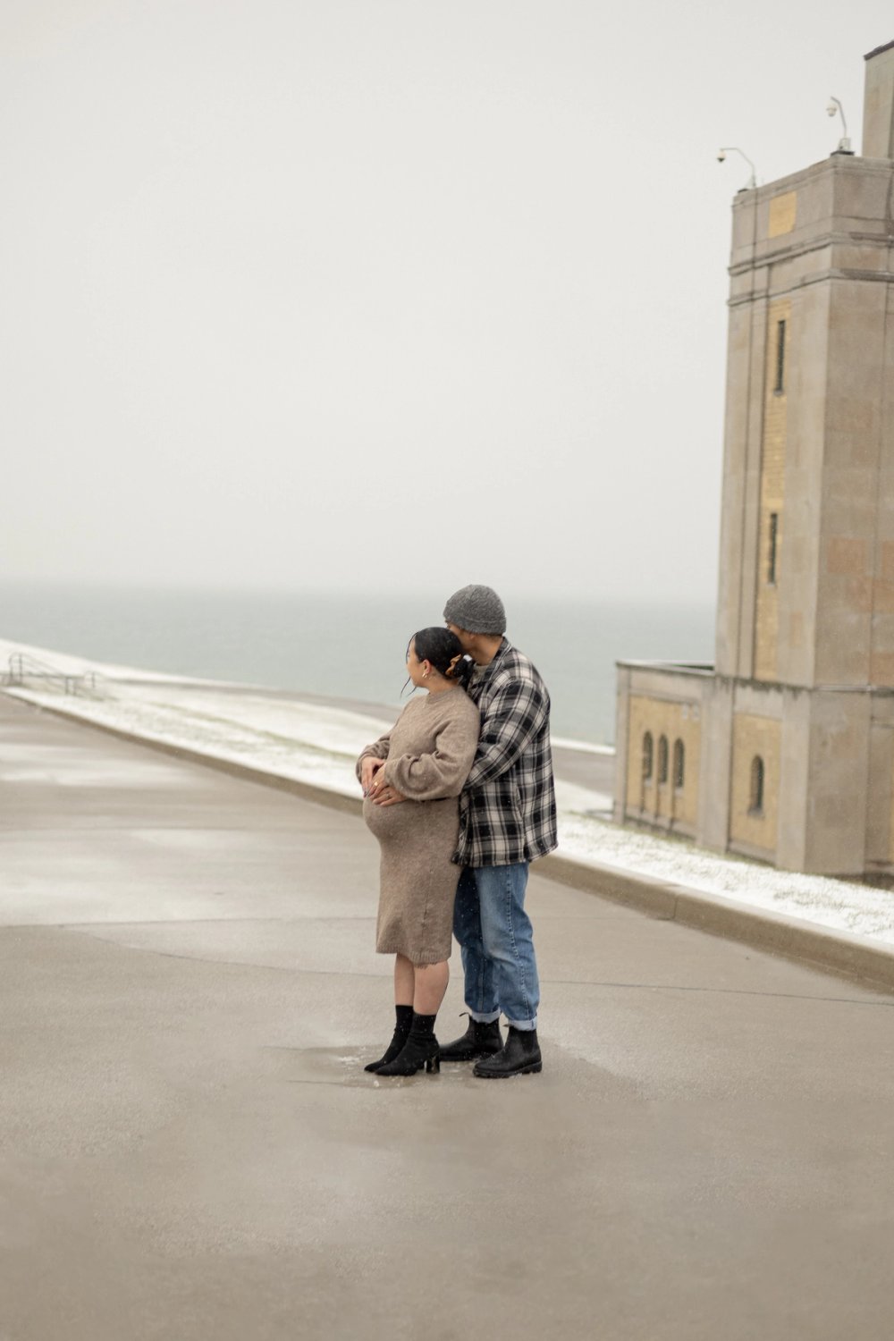Winter maternity photos at R.C. Harris Water Treatment Plant in Scarborough, ON. Dad hugging mom from behind holding baby bump while looking out at the lake - Newmarket Maternity Photographer
