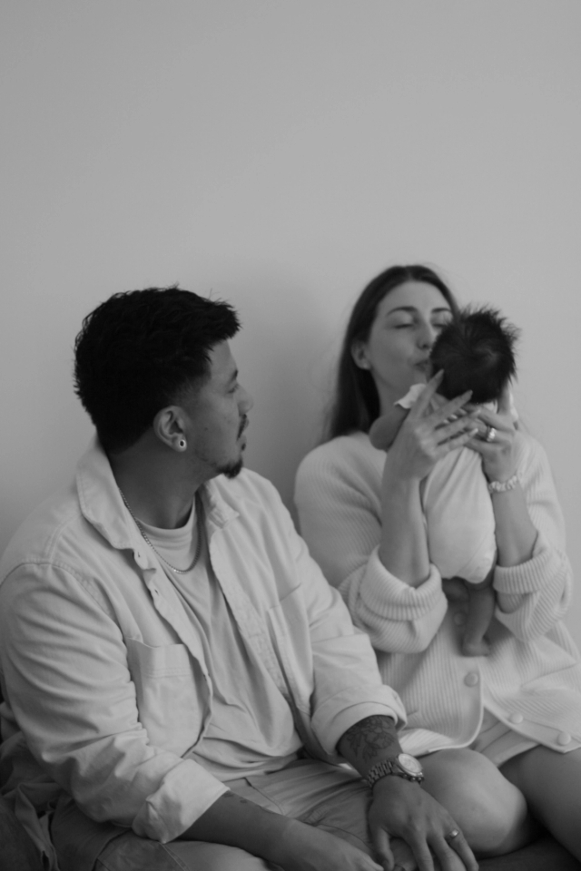 parents sitting on bench, dad is watching mom holding newborn baby upright in front of her, black & white - Newborn Photography