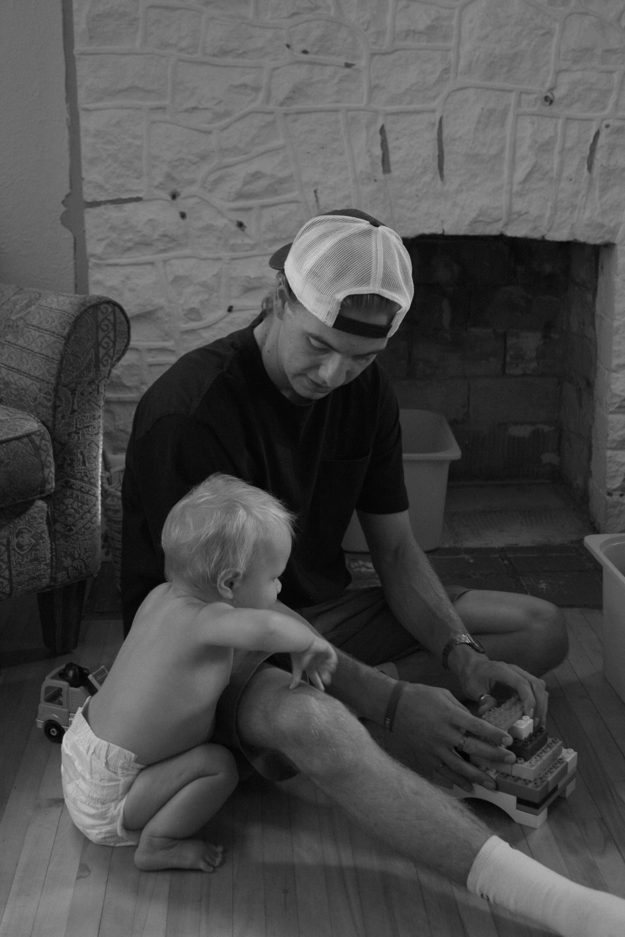 black and white image of baby and dad sitting on floor in front of fireplace, baby is leaning on dad's leg while dad builds lego - Family Photography