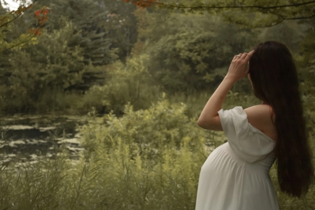 mom in white flowing dress and long hair holding hand up to face looking out at a green pond - Maternity Photography