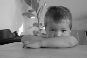 black and white image of Toddler sitting on chair with arms crossed on table, peaking slightly over arms toward the camera - Family Photography