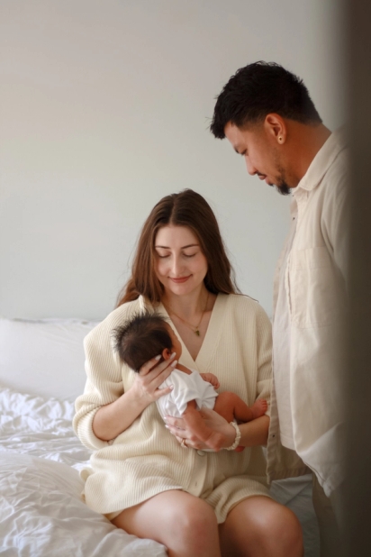 Mother sitting on bed craddling newborn baby and smiling while dad stands next to them looking down at the baby - Newborn Photography