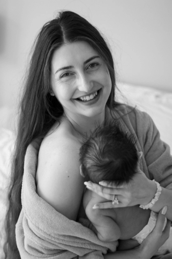 black and white photo of mom smiling at camera with robe off one shoulder, holding newborn baby facing away from camera - Newborn Photography