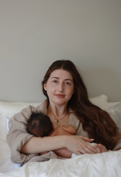 Mom sitting on bed breastfeeding newborn baby while looking at camera - In-home Newborn photography