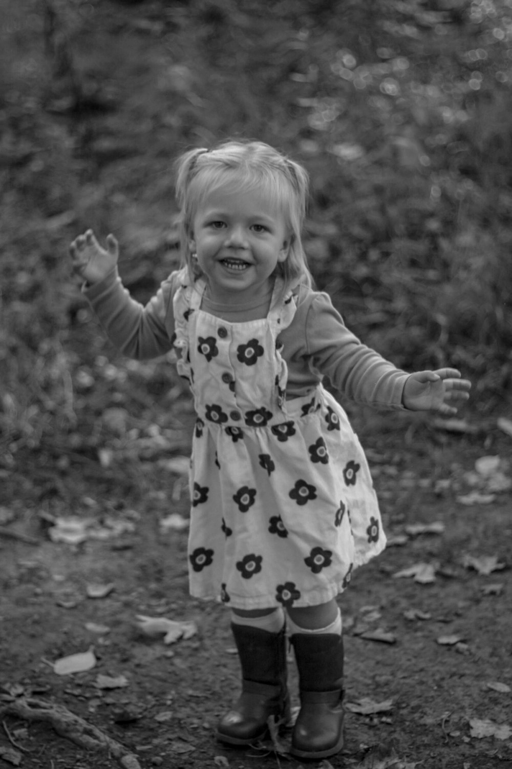 black and white photo of toddler girl in a floral dress and boots smiling at the camera with her hands out to the side -Newmarket Family Photography