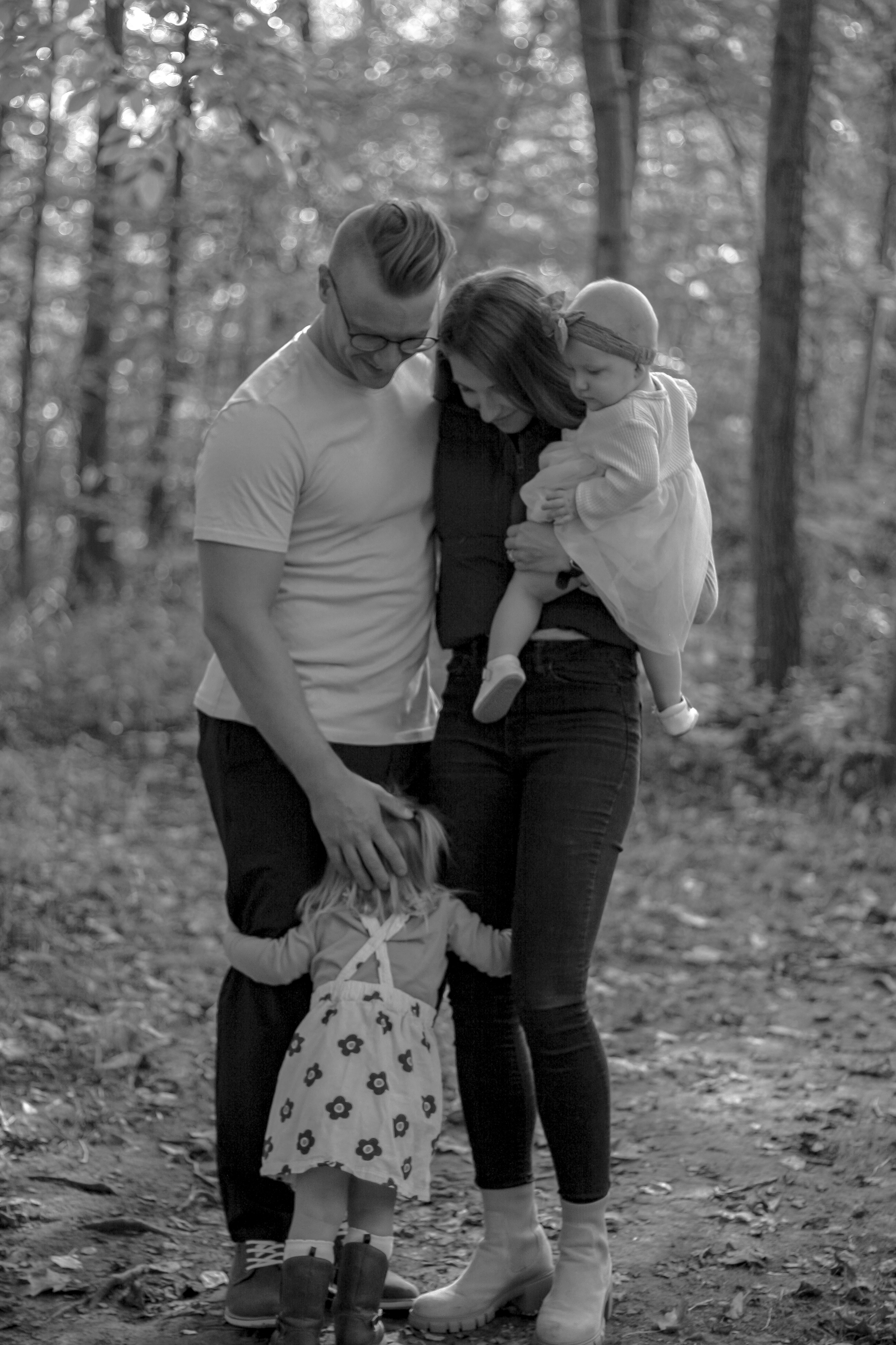 Black and white photo of family, mom is holding baby girl and toddler in floral dress is hugging mom and dad's legs, in middle of forest - Family Photography