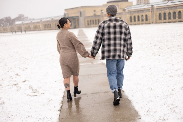 Candid moment of pregnant woman and her husband holding hands walking on a path surrounded by a light, airy snowfall.