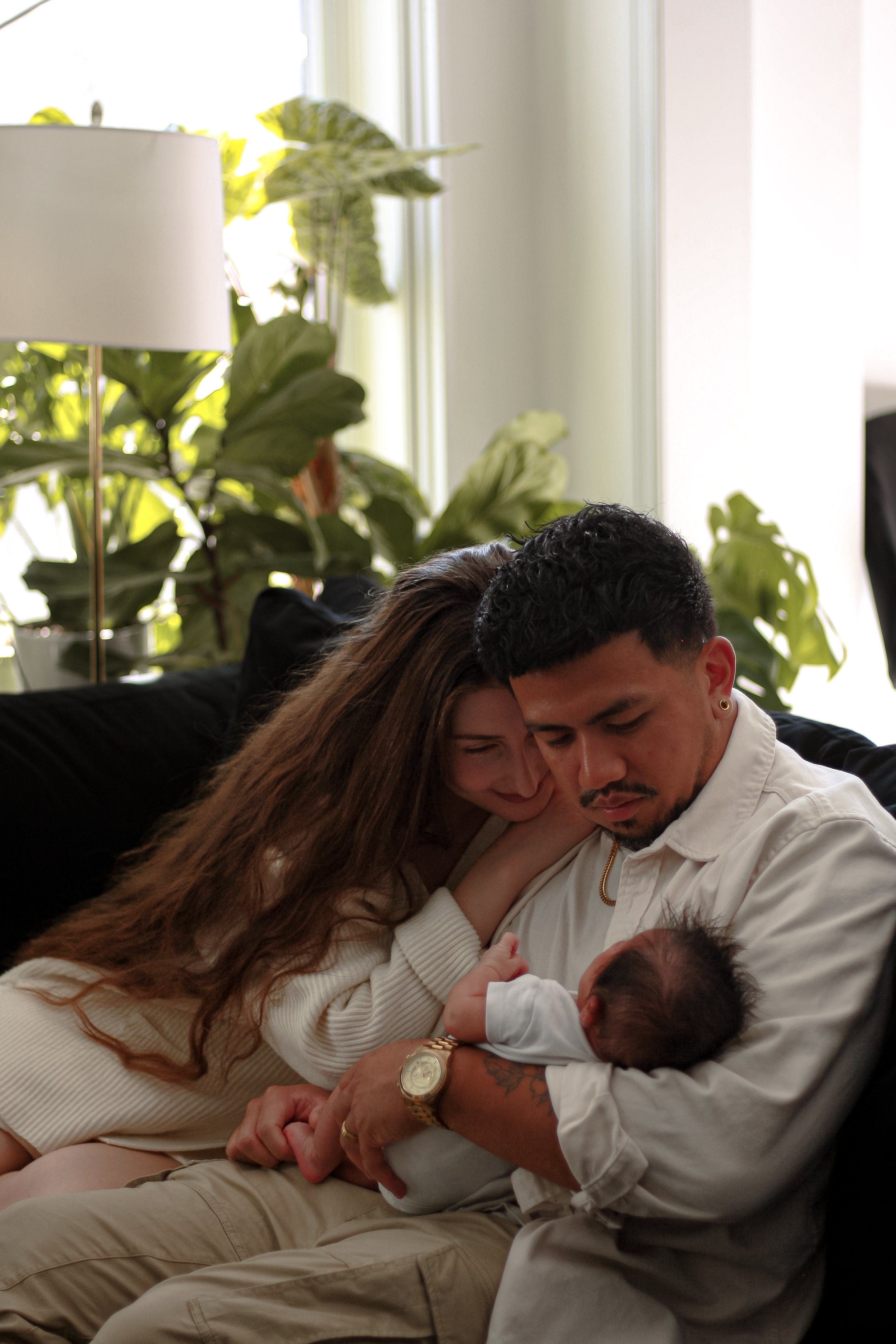 Couple sitting on couch, dad is craddling baby in arms while mom leans on dads shoulder both looking at the baby with green plants and a lamp in the background - Newmarket Newborn Photography