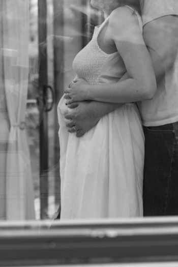 black and white photo of mom and dad holding baby bump view through window - maternity photos Newmarket