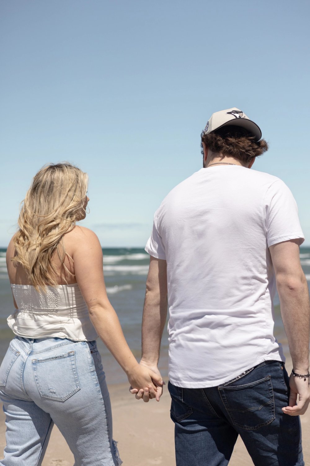 man and woman holding hands walking by the beach with their backs towards camera - Wasaga Beach Engagement Photography