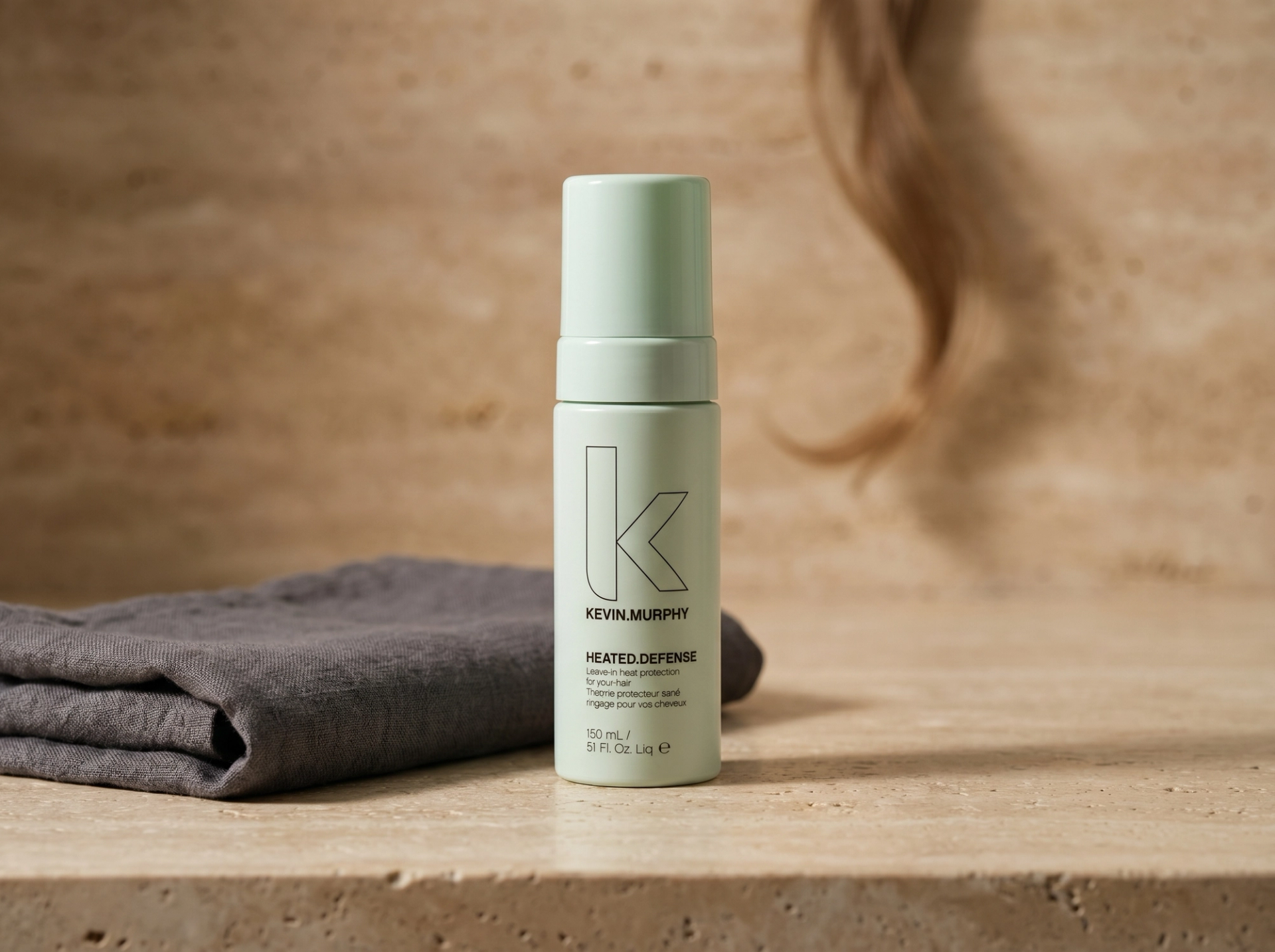 Editorial Scene: Kevin Murphy HEATED.DEFENSE auf warmem Travertin-Vanity mit gefaltetem charcoal-grauen Leinen-Handtuch und dunkelblondem Haarstrang im Bokeh – Australian-Spa-Mood