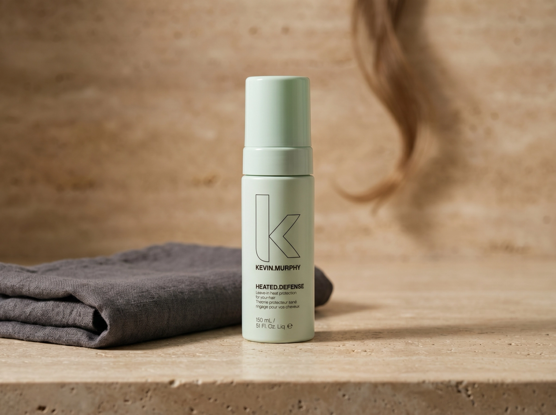 Editorial Scene: Kevin Murphy HEATED.DEFENSE auf warmem Travertin-Vanity mit gefaltetem charcoal-grauen Leinen-Handtuch und dunkelblondem Haarstrang im Bokeh – Australian-Spa-Mood