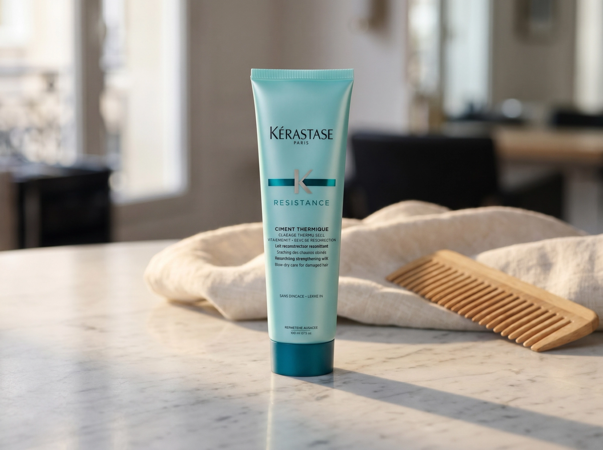 Editorial Scene: Kérastase Resistance Ciment Thermique Tube stehend auf Carrara-Marmor-Counter in einem Pariser Premium-Salon, mit gefaltetem cream-linen Tuch und natürlichem Wide-Tooth-Holzkamm im Bokeh, warmes Morgenlicht von links – Vogue-Editorial-Mockup