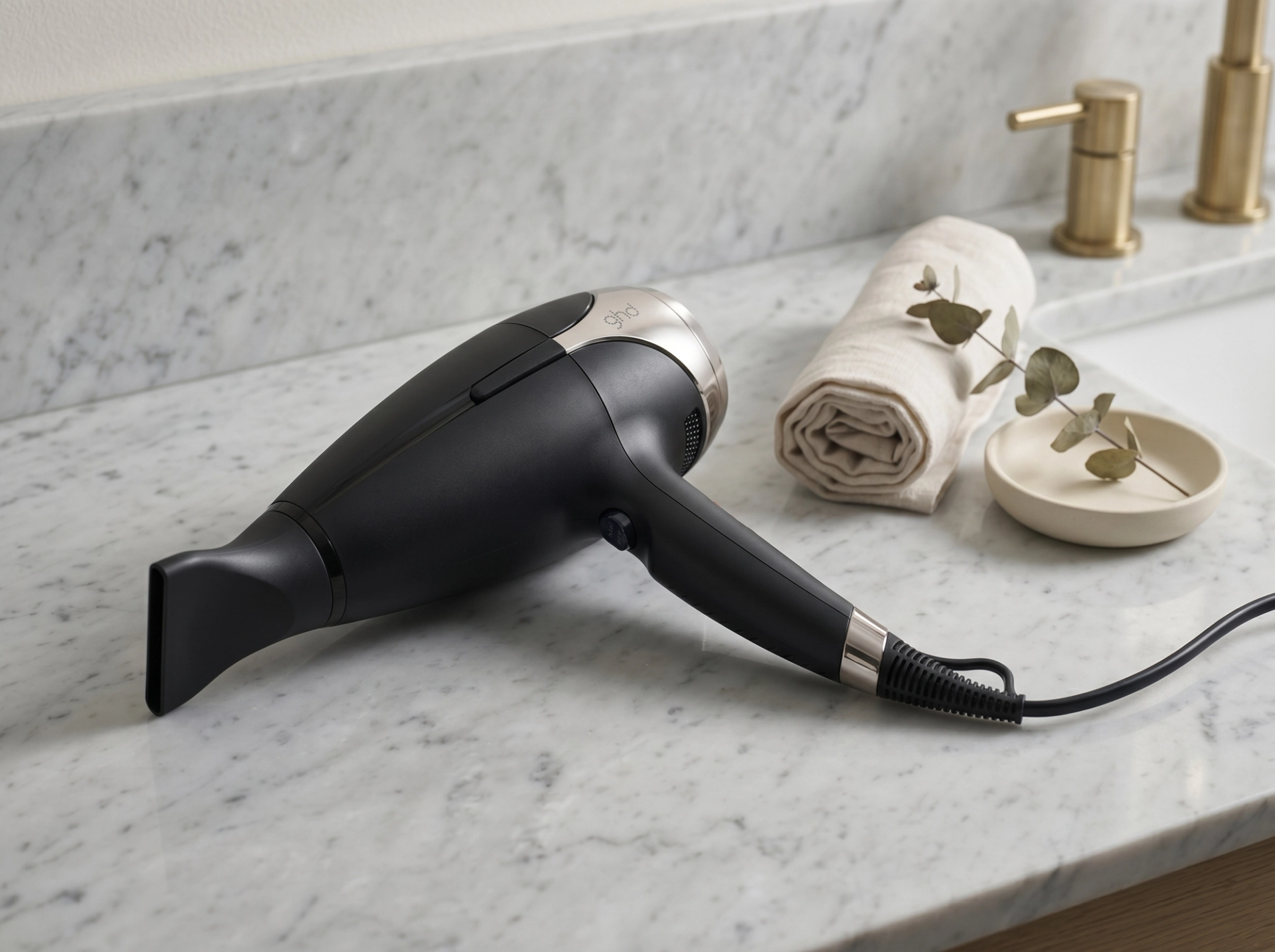 ghd Helios Föhn liegt flach auf einer hellen Carrara-Marmor-Salon-Vanity neben einer gerollten Leinen-Serviette und einer Keramikschale mit Eukalyptus
