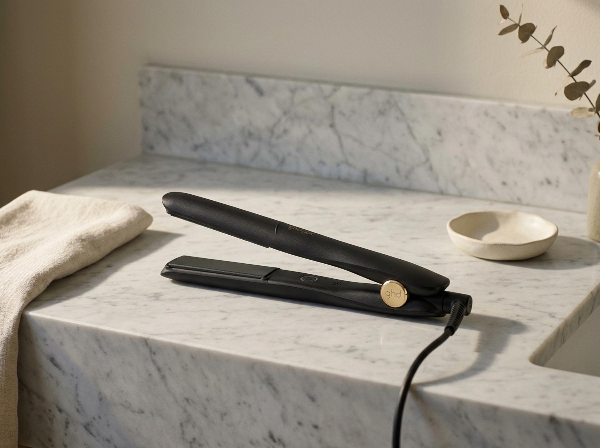ghd Gold Styler Glätteisen liegt flach auf einem hellen Marmor-Waschtisch mit gefaltetem cremefarbenem Leinen-Handtuch und einer matten Keramikschale