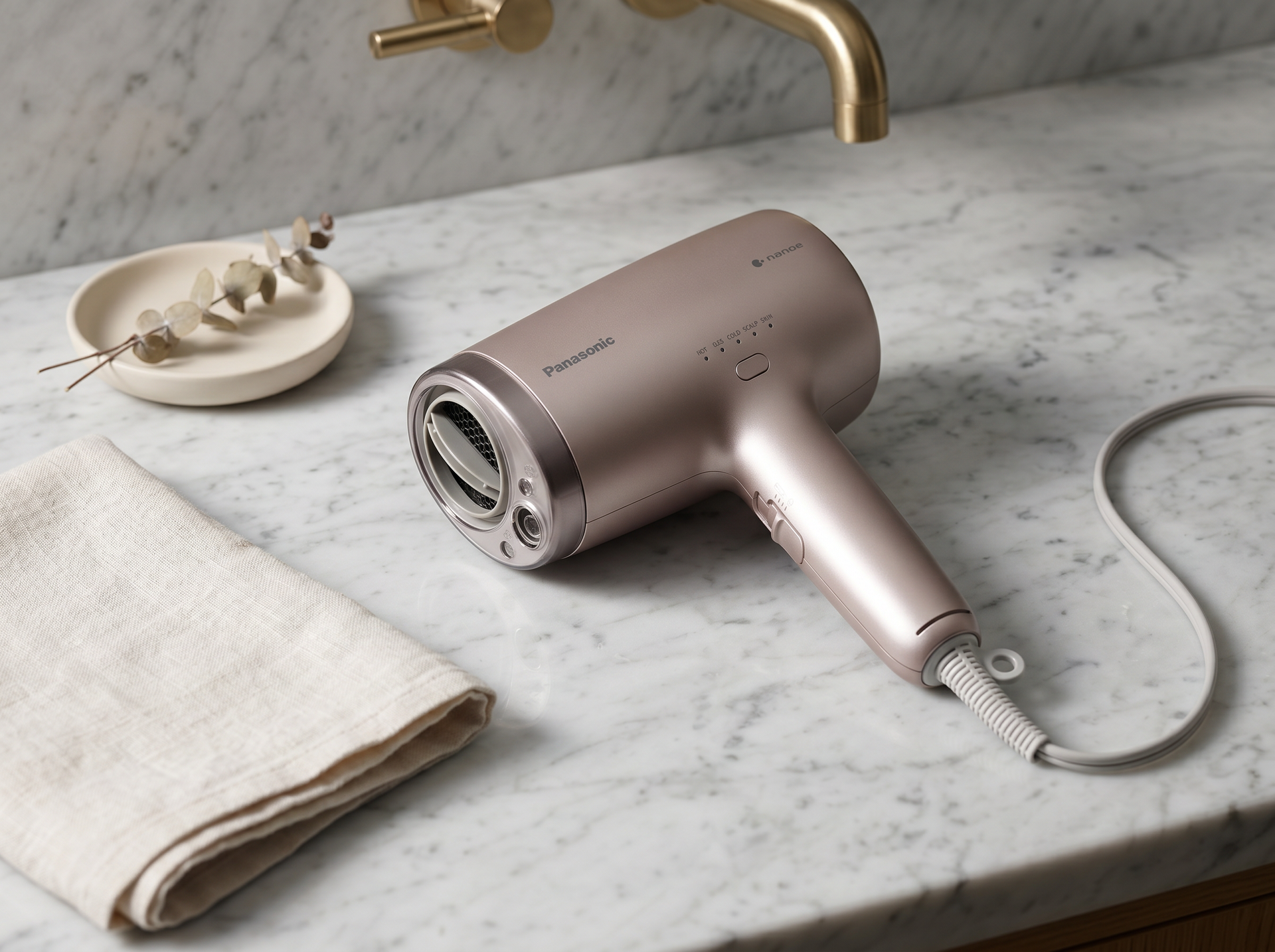 Editorial Bathroom-Vanity-Scene: Panasonic EH-NA9N liegt flach auf hellem Carrara-Marmor neben gefaltetem Cream-Linen-Tuch und Eukalyptus-Schale, brushed-brass Wasserhahn im Bokeh – Premium Salon-Mockup