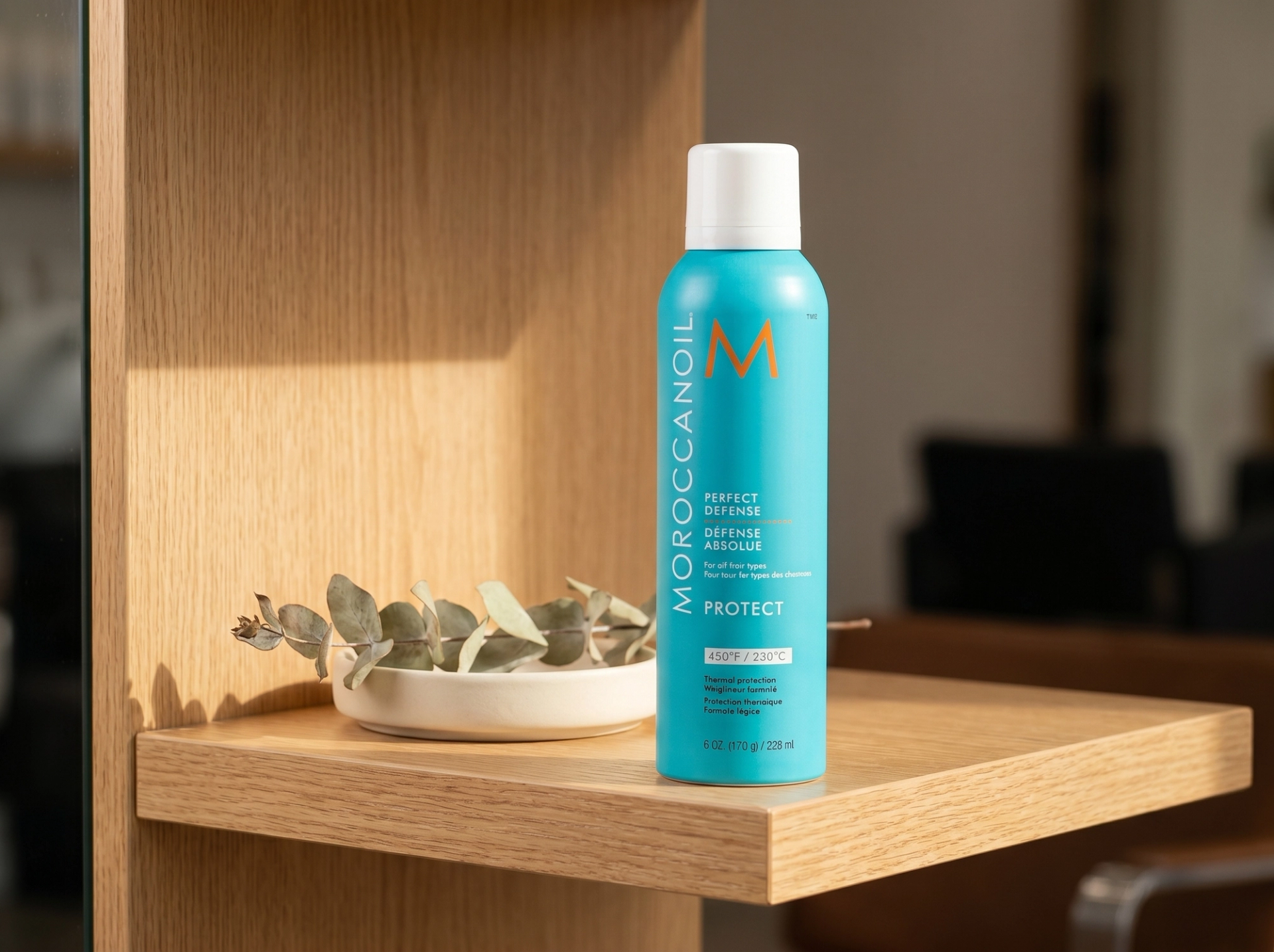 Editorial Scene: Moroccanoil Perfect Defense Aerosol-Dose stehend auf warmem Light-Oak-Salon-Shelf, daneben getrocknete Eukalyptus-Stalken in matte-cream Keramikschale, warmes Morgenlicht – Premium-Mockup