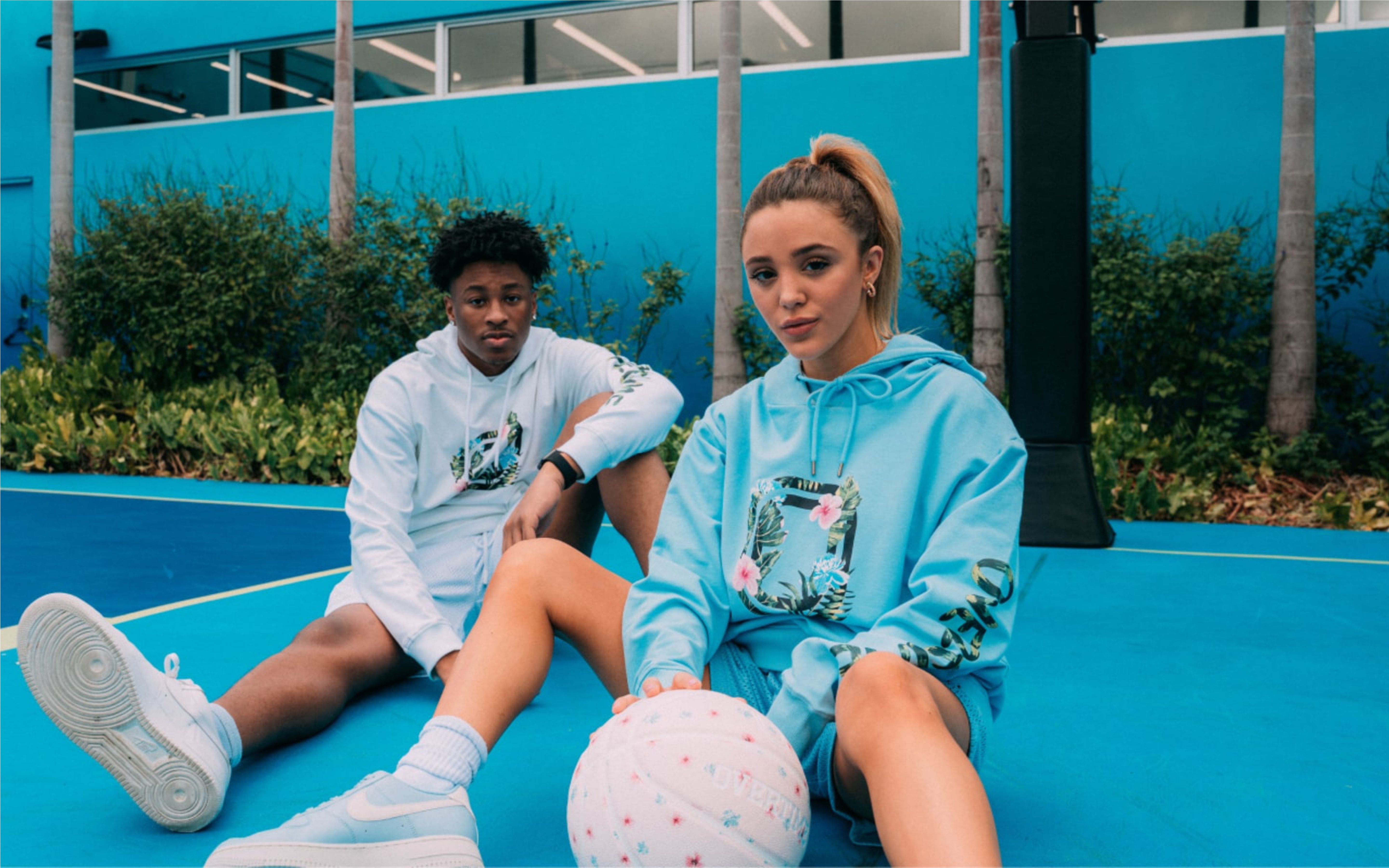 Two teens sit on a basketball court wearing Overtime hoodies