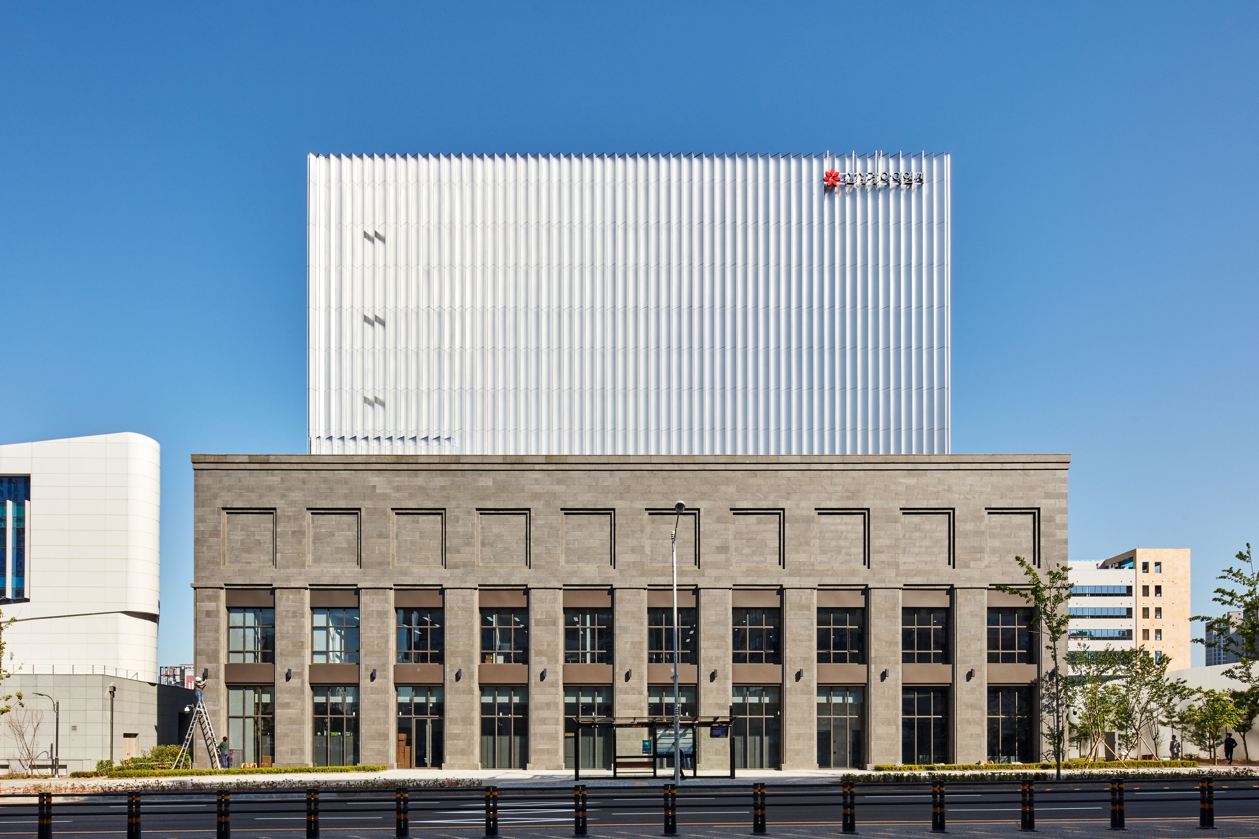 A front-facing daytime photograph of the Shinsegae I&C Data Center building. The structure has a two‑part design: a lower section with a stone-like exterior featuring tall rectangular windows and architectural detailing, and an upper section with a sleek, modern white façade composed of vertical ribbed panels. The Shinsegae I&C logo appears on the upper portion. The sky is clear and bright, creating a clean, minimal backdrop for the building.
