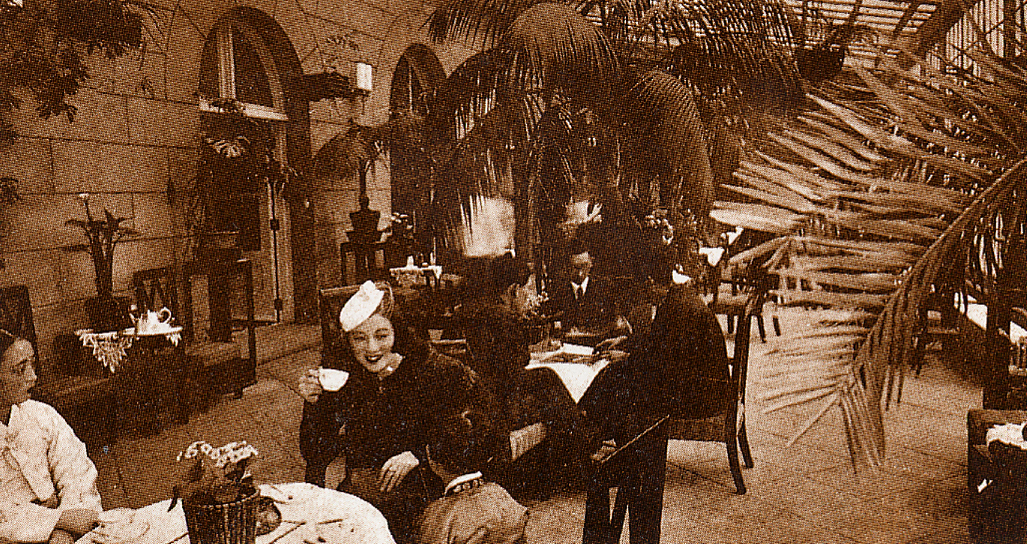 An archival sepia-toned photograph of an outdoor café area at the Chosen Hotel. Several people sit at small round tables drinking tea or coffee. Potted plants and tall palm fronds surround the seating area, creating a garden-like atmosphere. Behind the tables, arched doorways and stone walls of the historic hotel building are visible. The scene captures a leisurely social moment from the early 20th century.