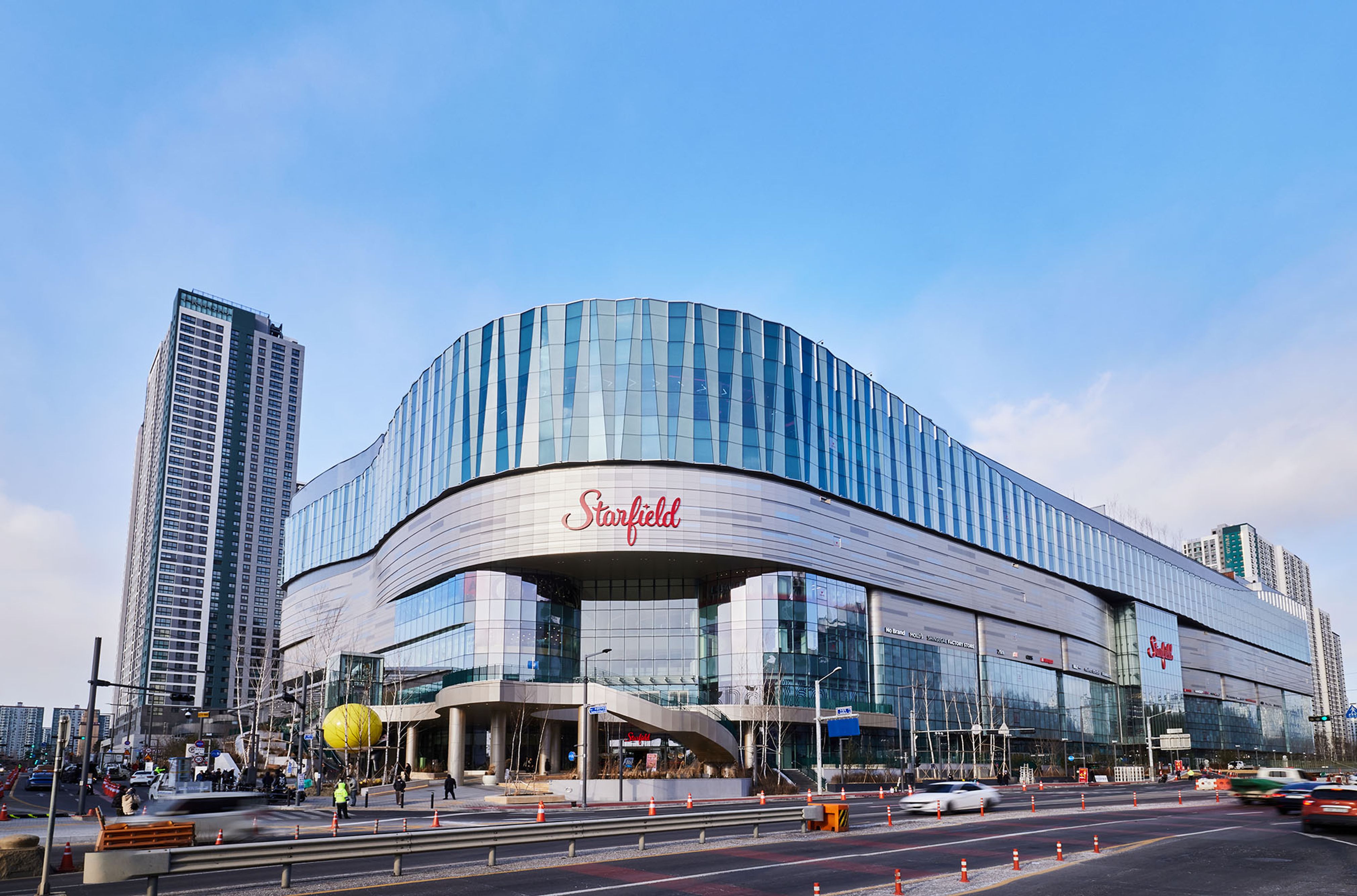 A daytime view of Starfield Suwon, a large modern shopping complex with a sweeping curved façade made of reflective blue‑tinted glass panels. The building’s ‘Starfield’ logo is displayed prominently near the center. Wide entrance steps and glass doors lead into the mall, with cars and pedestrians visible along the road in front. High‑rise buildings surround the structure under a bright, lightly clouded sky.