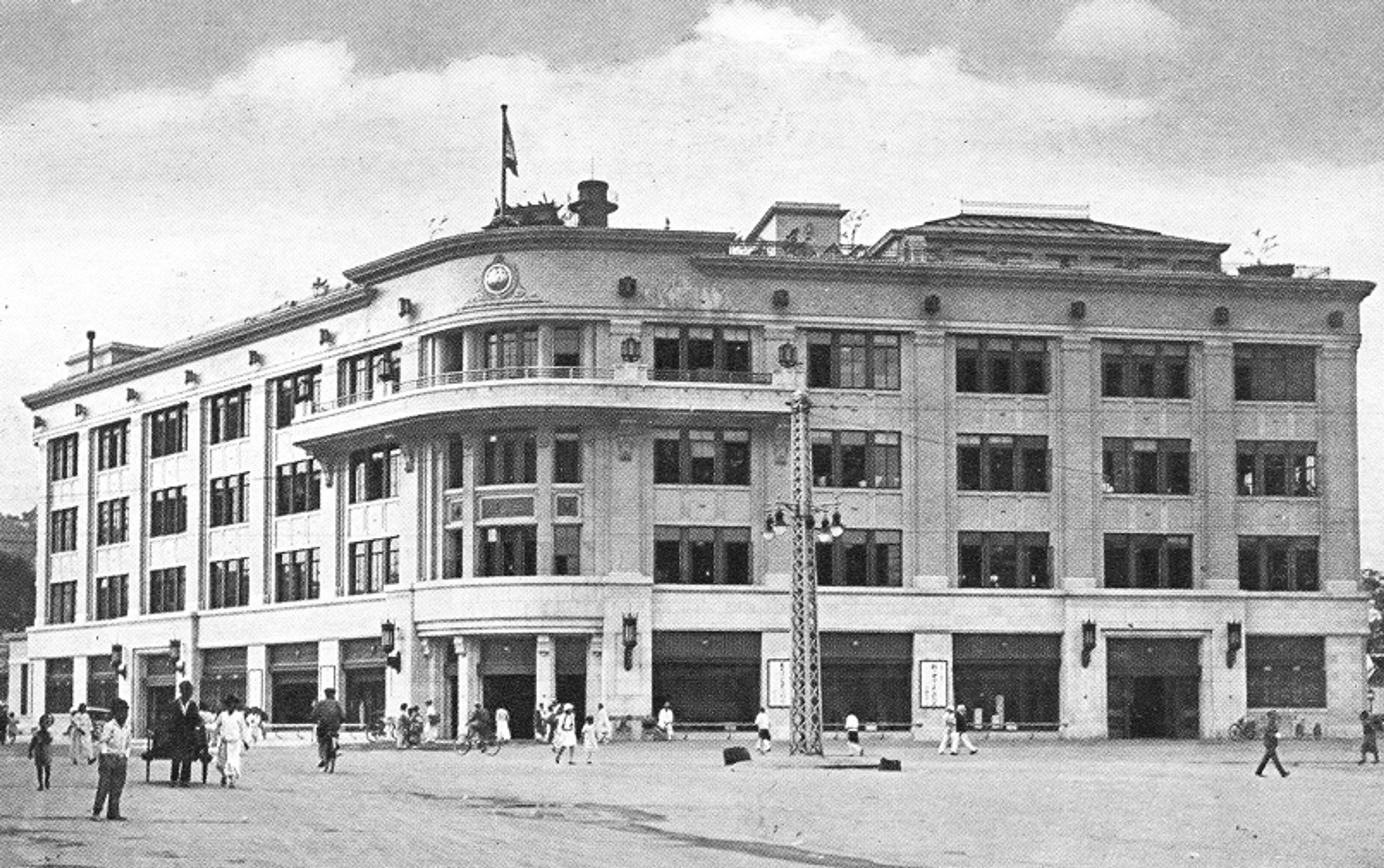 An archival black‑and‑white photograph of the Mitsukoshi Department Store’s Seoul branch, the historic predecessor of today’s Shinsegae flagship. The large, multi‑story building features a curved corner façade, rows of rectangular windows, and decorative architectural details. A flag flies from the rooftop. In the open plaza in front of the building, people walk across the wide, empty space, some in traditional attire and others in early 20th‑century Western clothing. The scene conveys the scale and prominence of the department store during that era.
