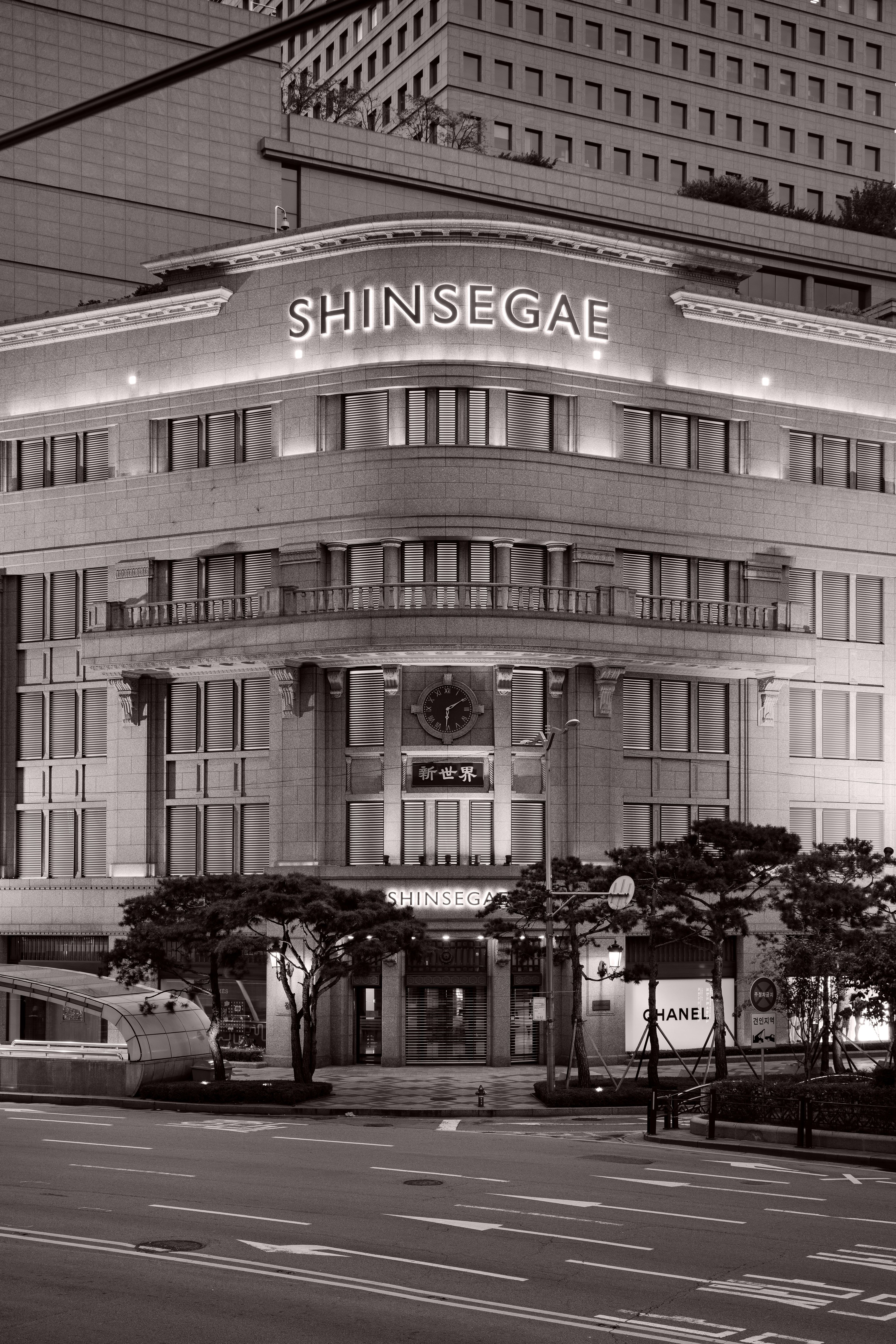A nighttime street view of a large, historic-style department store building with the illuminated sign ‘Shinsegae’ at the top. The facade features rounded corners, tall windows, and a central entrance marked by a clock above the doorway. Trees, streetlights, and a mostly empty road are visible in the foreground, with modern high‑rise buildings in the background.