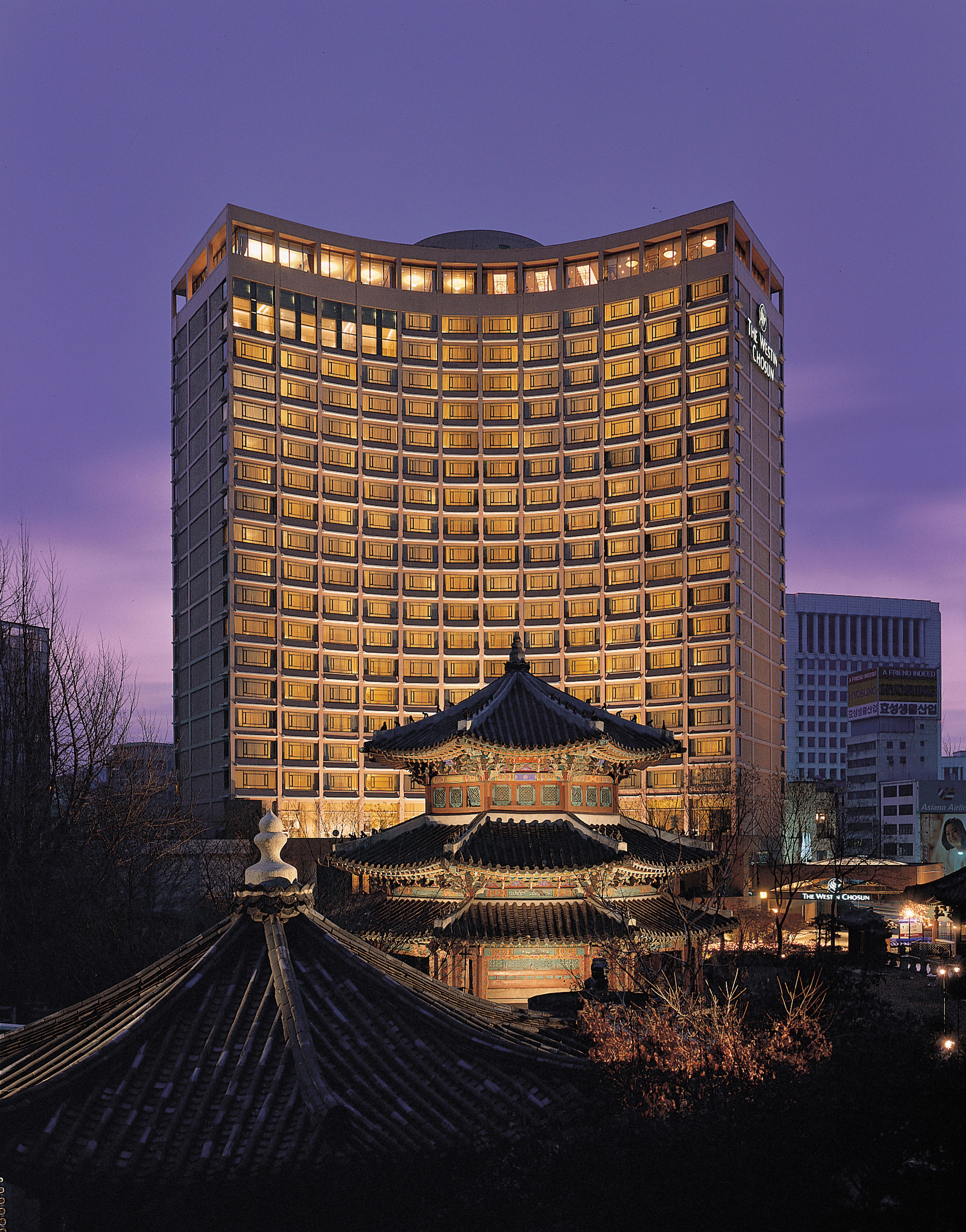 A twilight view of the Westin Josun Seoul hotel, with its tall, gently curved façade illuminated by warm window lights against a purple sky. In the foreground stands a traditional Korean pavilion with layered, tiled roofs, creating a striking contrast between historic architecture and the modern hotel tower behind it.