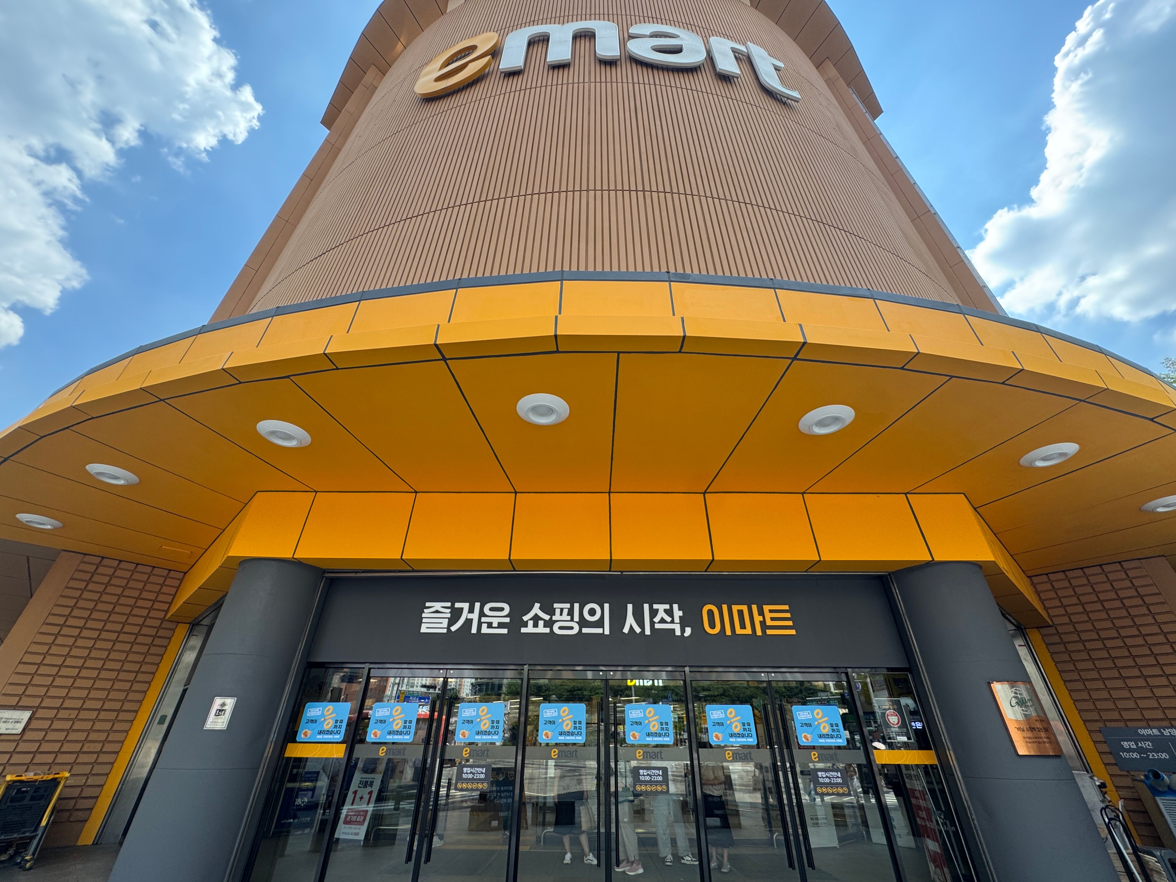 A low‑angle daytime photograph of the exterior entrance of an emart store. The building features a tall, rounded façade with vertical textured panels and a large ‘emart’ logo near the top. A deep yellow canopy extends over the glass entrance doors, which display blue promotional posters. Above the doors, a black sign reads ‘즐거운 쇼핑의 시작, 이마트’ (‘Fun starts here, emart’). The sky is bright with a few scattered clouds.
