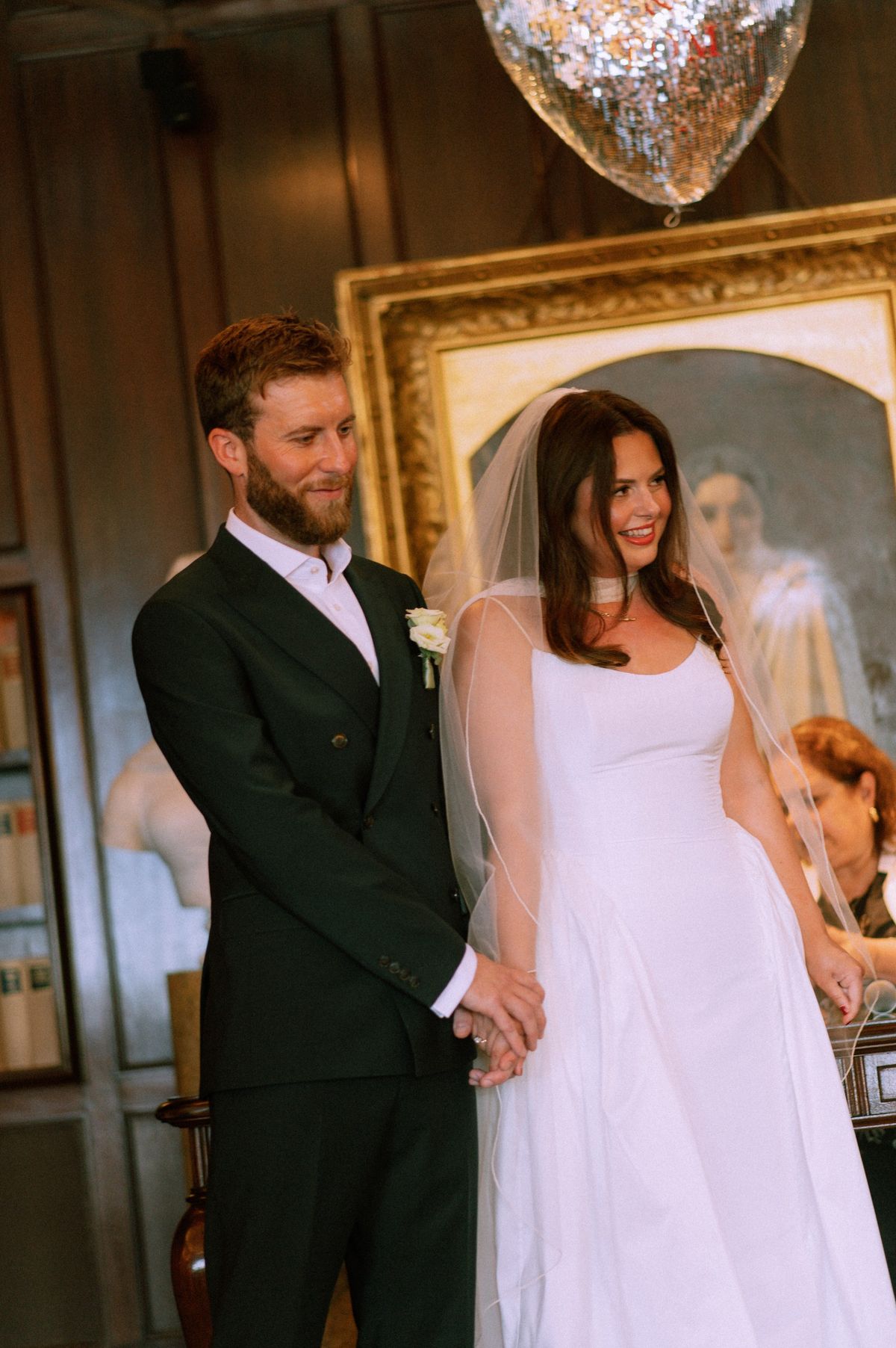 Bride and groom holding hands in a wood-panelled room beside a gilt-framed portrait