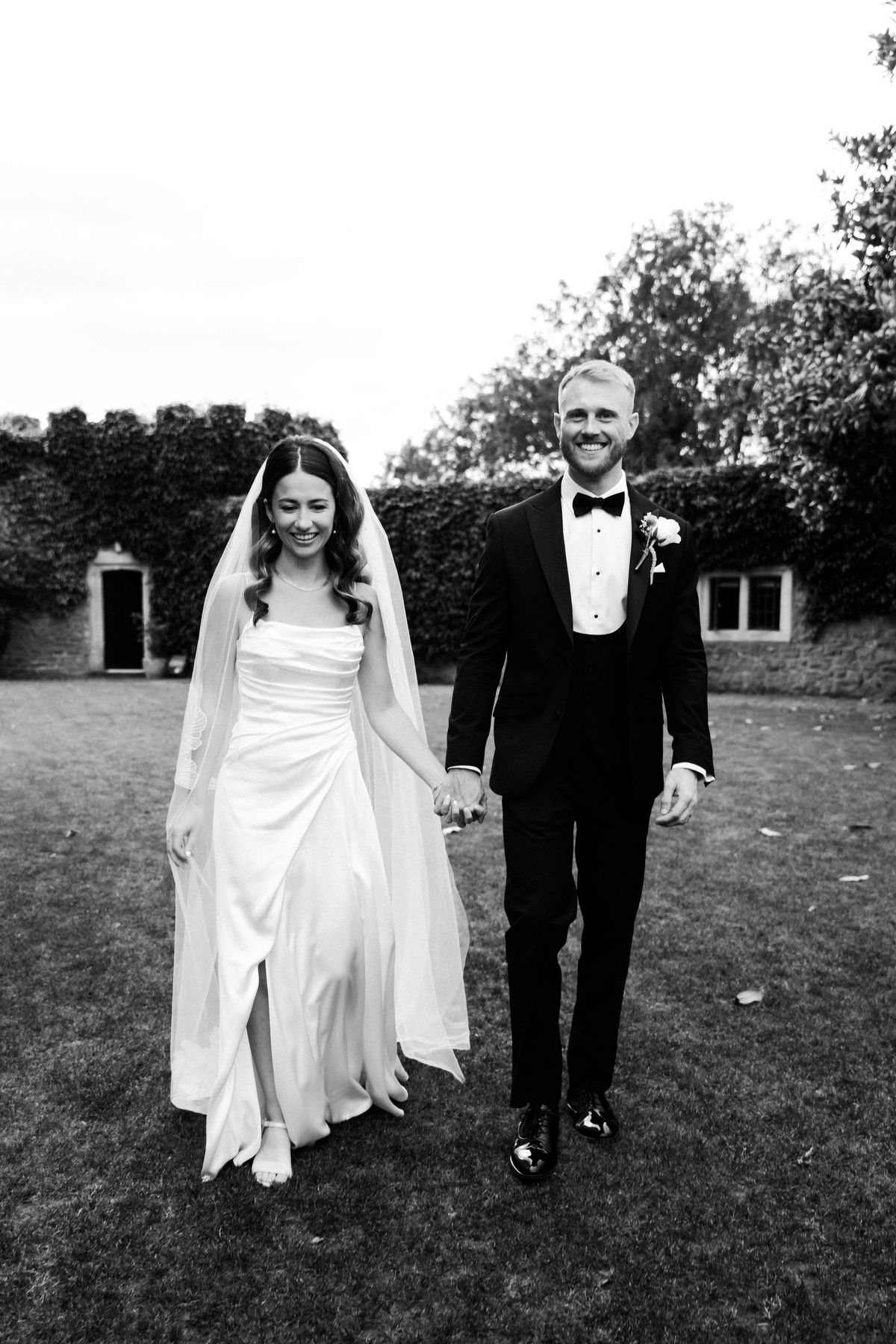 Black and white photo of a bride and groom walking hand in hand across a lawn in front of a castle wall