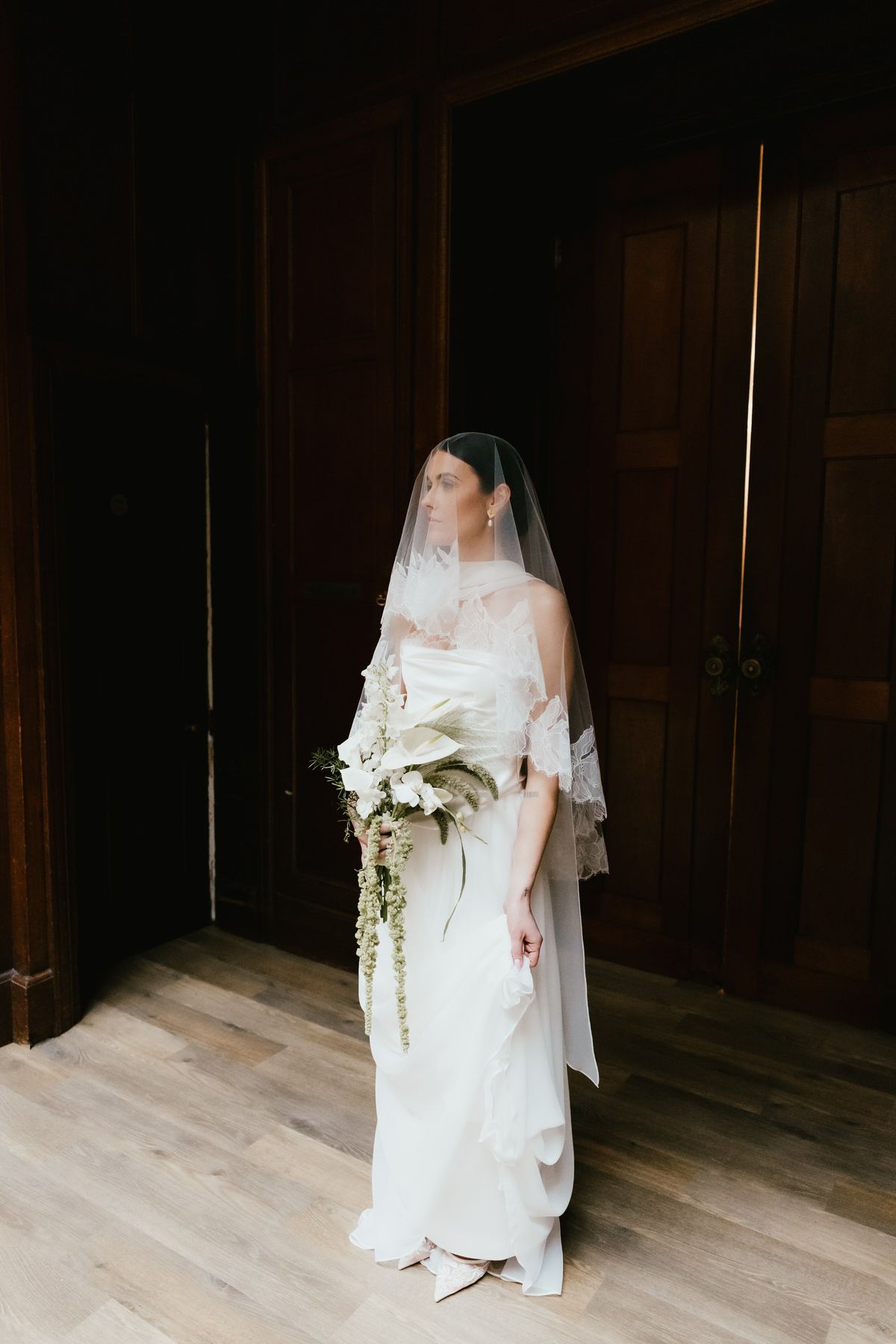 Bride in a blusher veil with lace edging, holding a cascading bouquet in a doorway