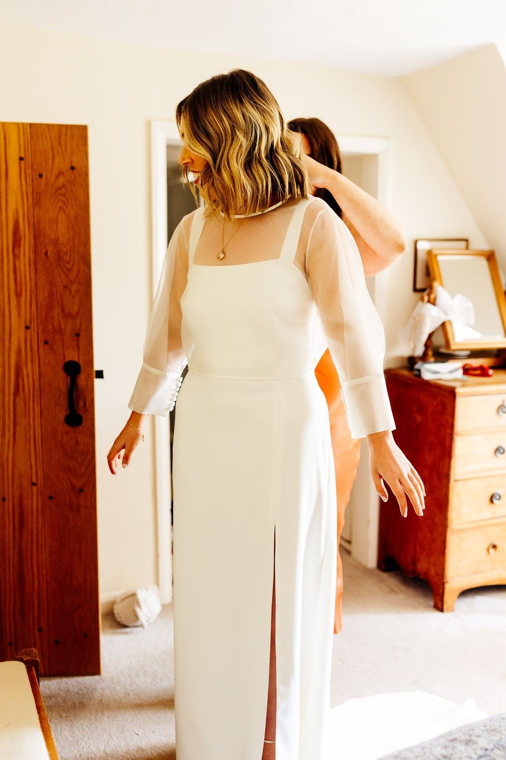 Bride in a sheer organza top and crepe dress adjusting her sleeve in a bedroom