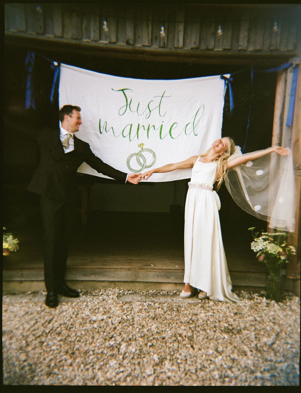 Film photograph of a couple holding a Just Married banner outside a barn, the bride in a vintage veil