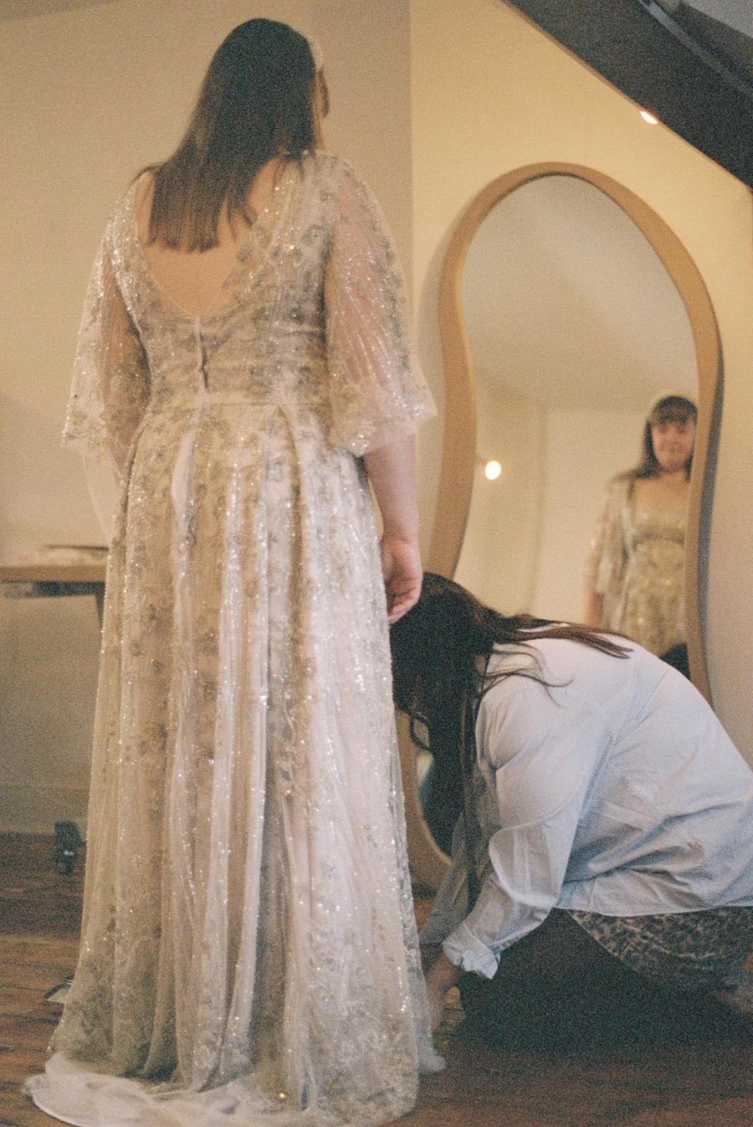 Bride seen from behind in an embroidered blush dress, looking into a mirror