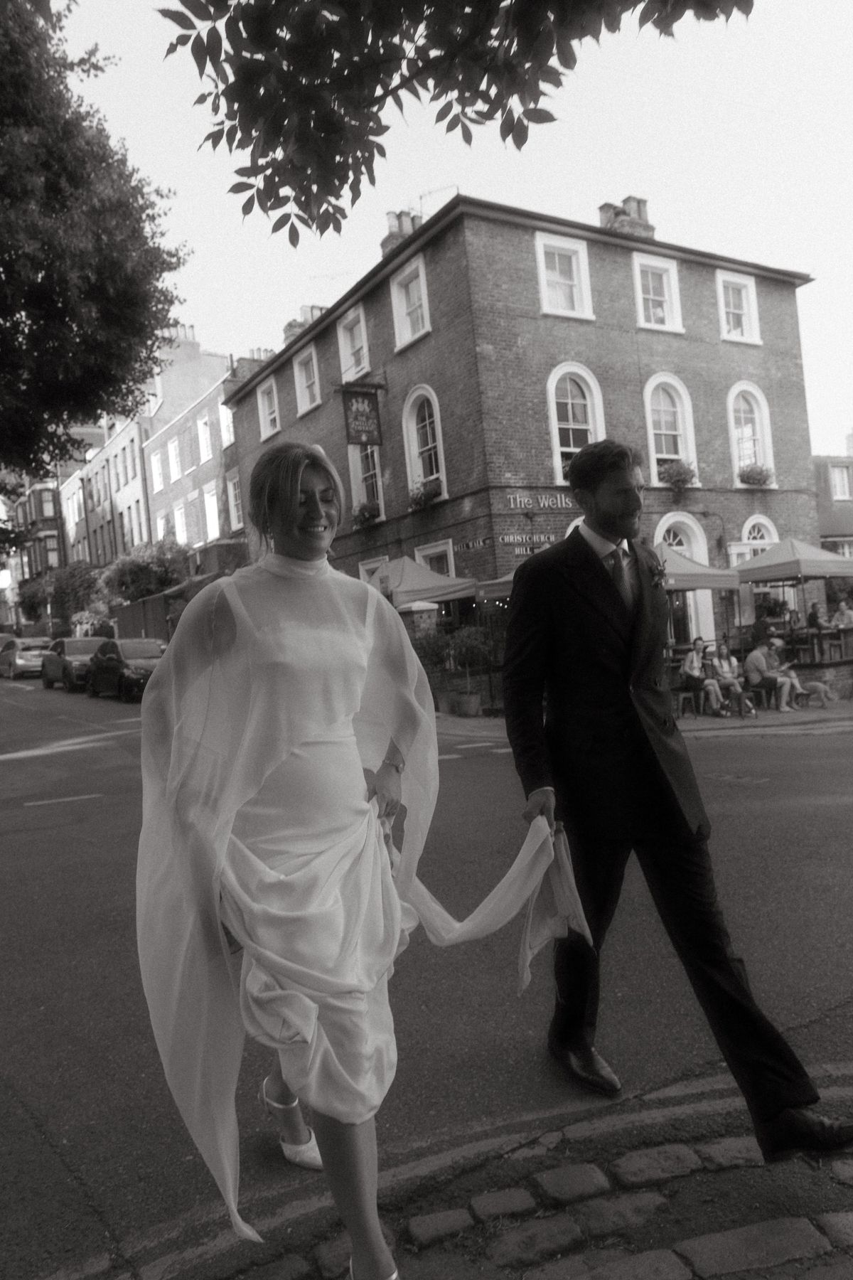 Bride in a chiffon cape walking hand in hand with her groom past Georgian houses