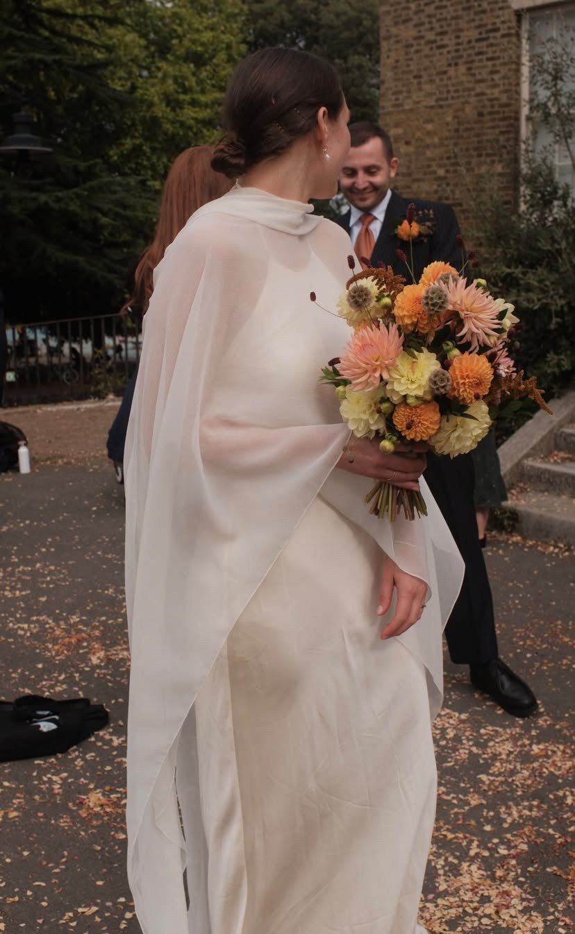 Bride in a silk chiffon cape holding an orange and yellow bouquet, her groom behind her