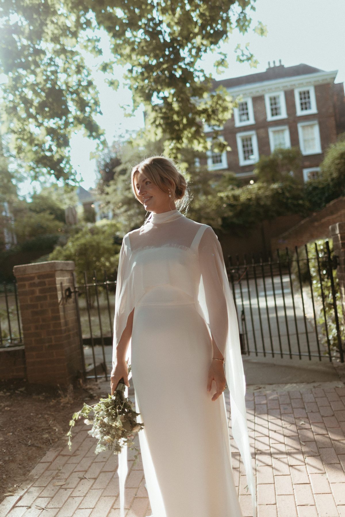 Bride in a chiffon cape and tailored crepe column dress by a gate on a sunny street