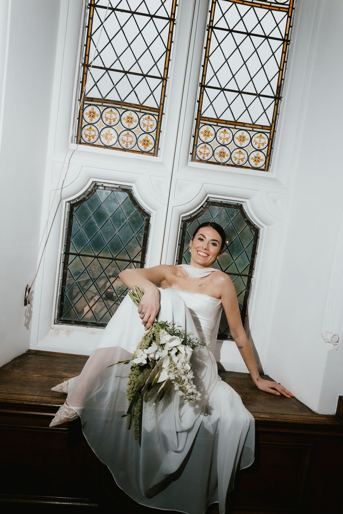 Bride seated on a window seat by stained glass windows, holding a bouquet