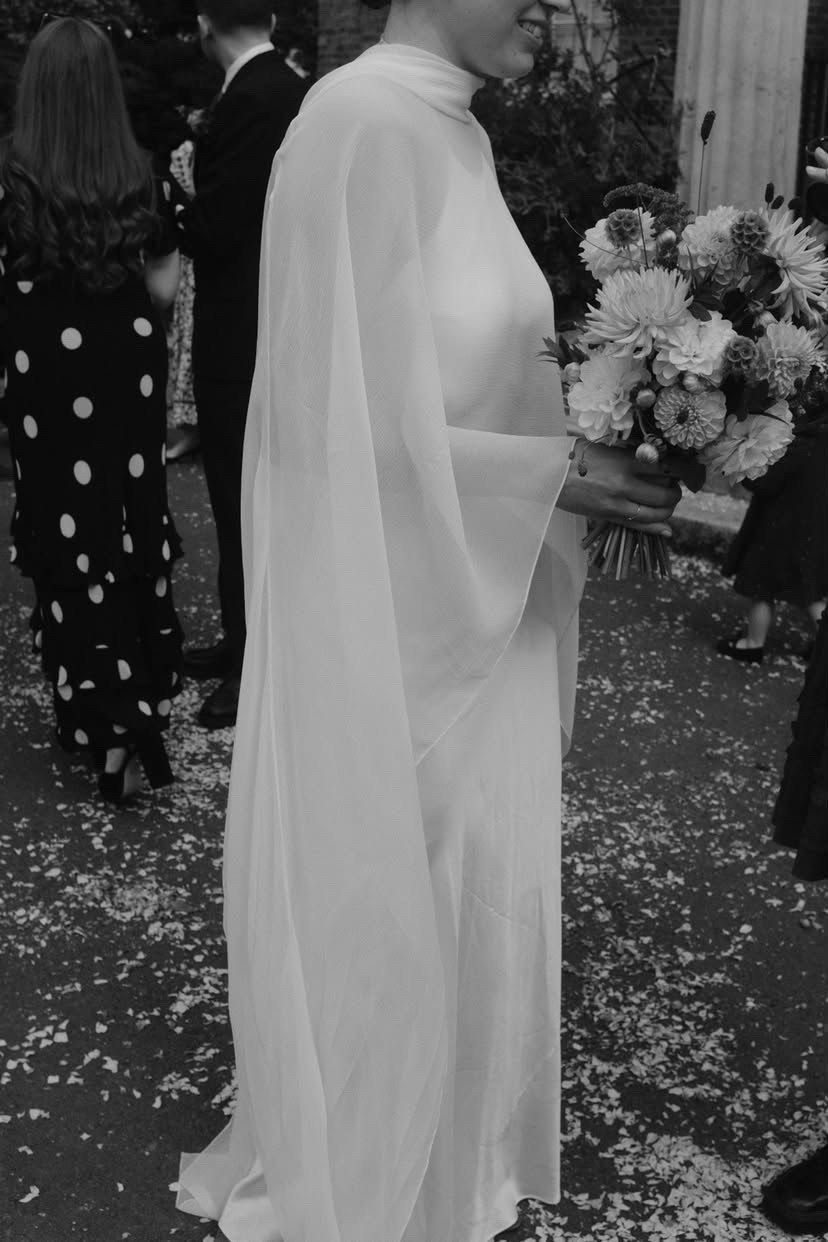 Black and white close-up of the flowing sleeves of a silk chiffon cape worn by a bride holding a bouquet