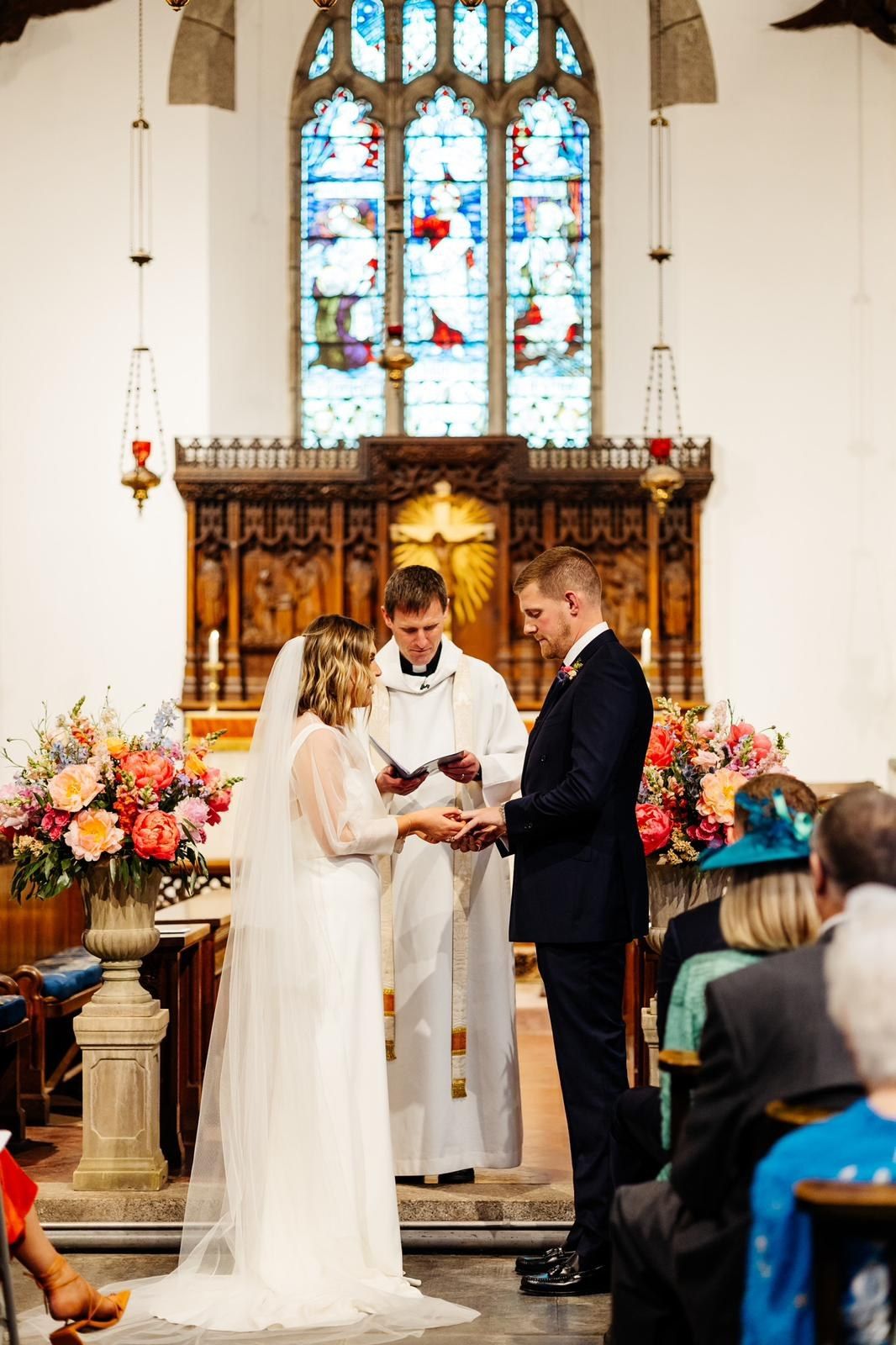 Couple exchanging rings at the altar of a church beneath a stained glass window