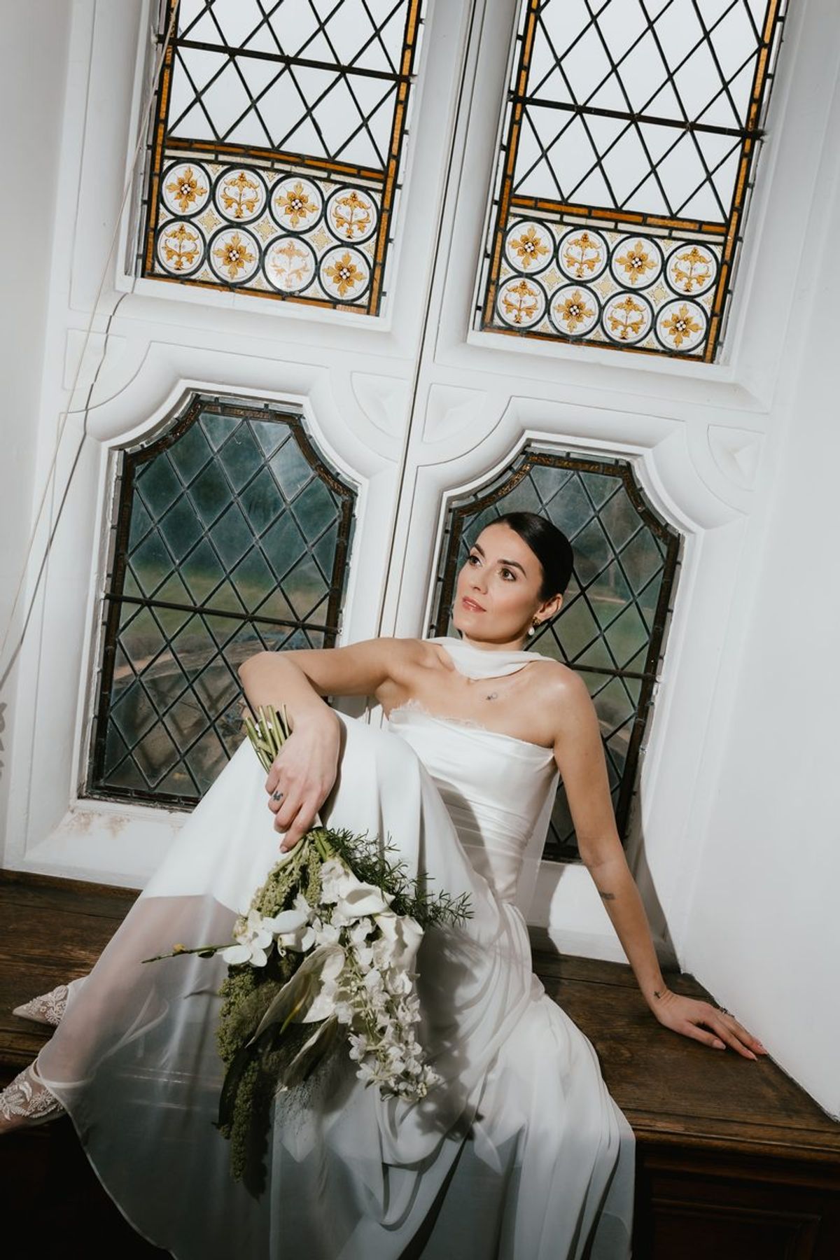 Bride seated by ornate stained glass windows holding bouquet