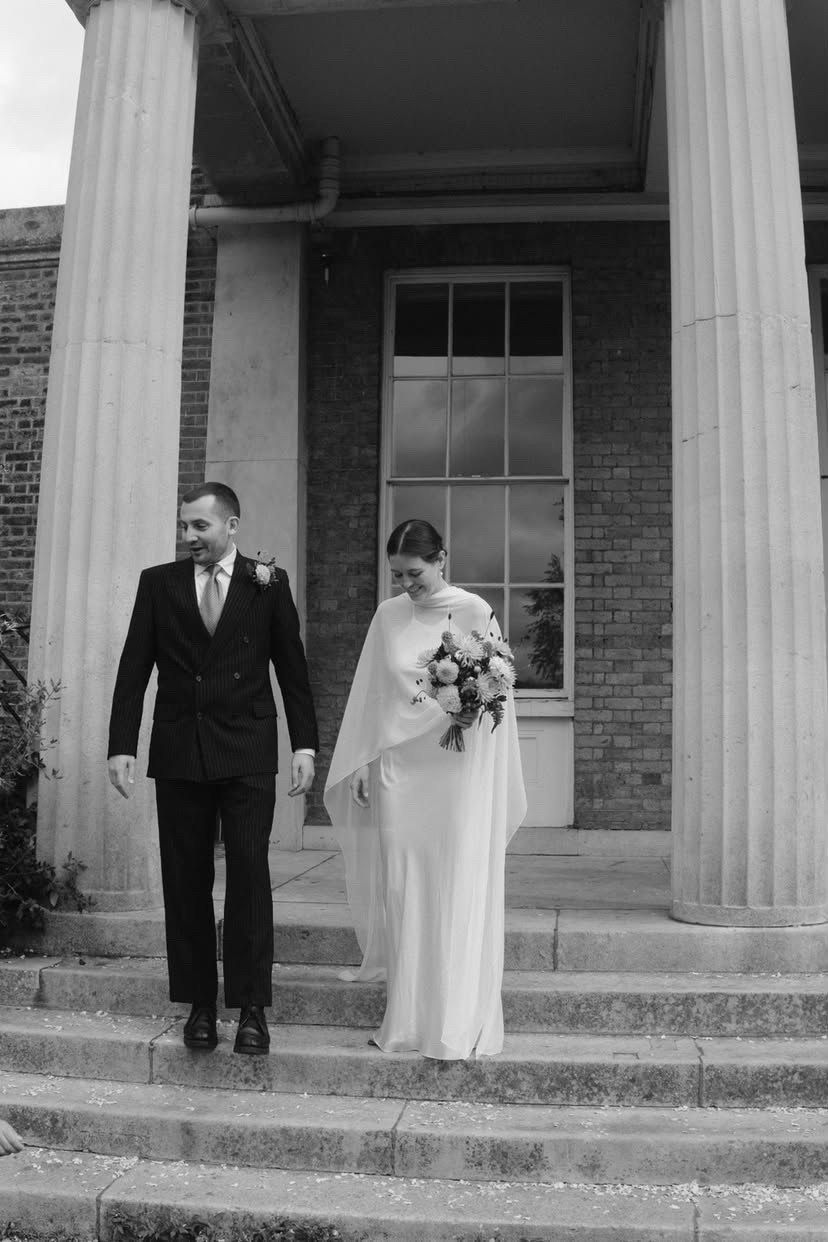 Black and white photo of a bride in a chiffon cape and her groom walking down steps between stone columns