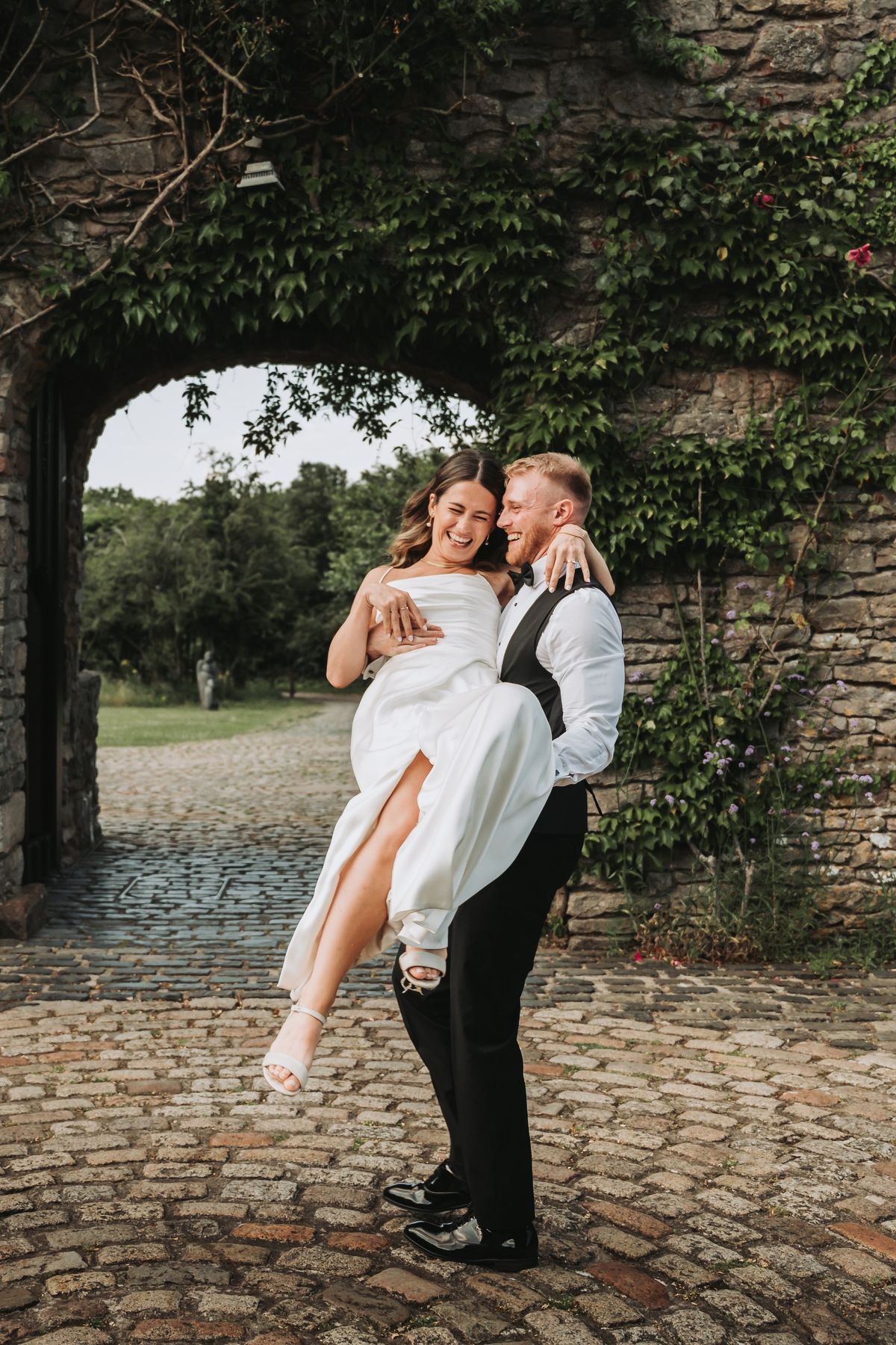 Groom lifting his bride in a draped satin dress beneath an ivy-covered stone arch